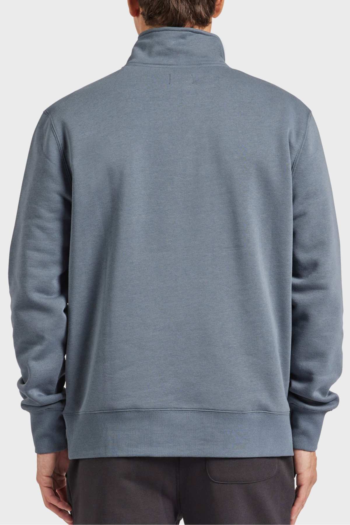 A person is shown from the back, dressed in an Academy Brand 1/2 Zip Sweat featuring a high collar and long sleeves in a grayish-blue hue, crafted from premium brushed fleece. The individual also appears to be wearing dark-colored pants against a plain white background.