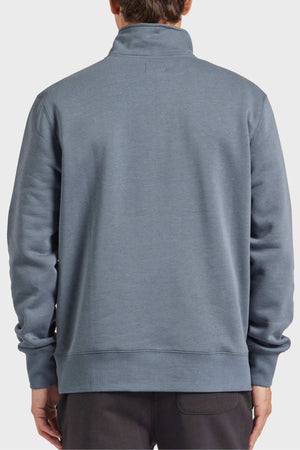 A person is shown from the back, dressed in an Academy Brand 1/2 Zip Sweat featuring a high collar and long sleeves in a grayish-blue hue, crafted from premium brushed fleece. The individual also appears to be wearing dark-colored pants against a plain white background.