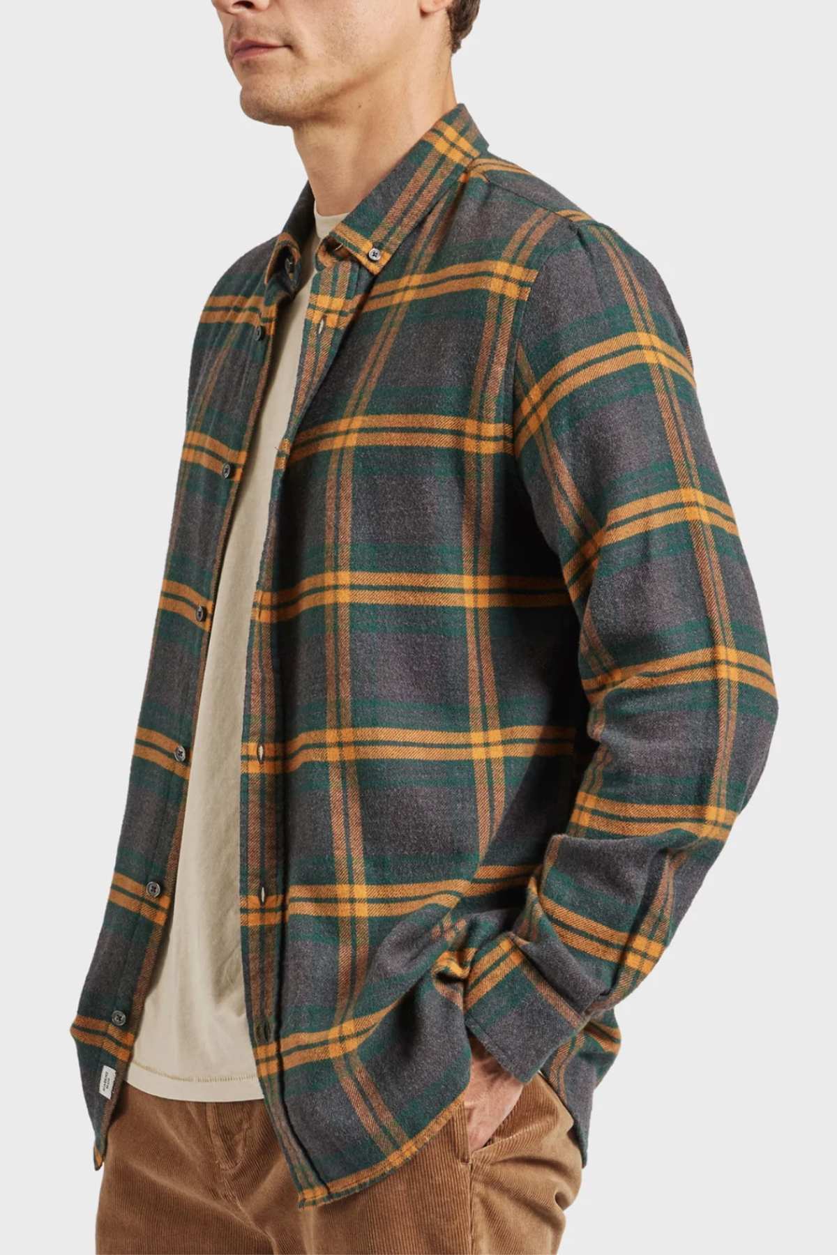 Wearing an Academy Brand Denver Shirt in green and orange plaid over a beige T-shirt, a person stands with hands in pockets. The cotton shirt is buttoned with long sleeves against a plain white background.