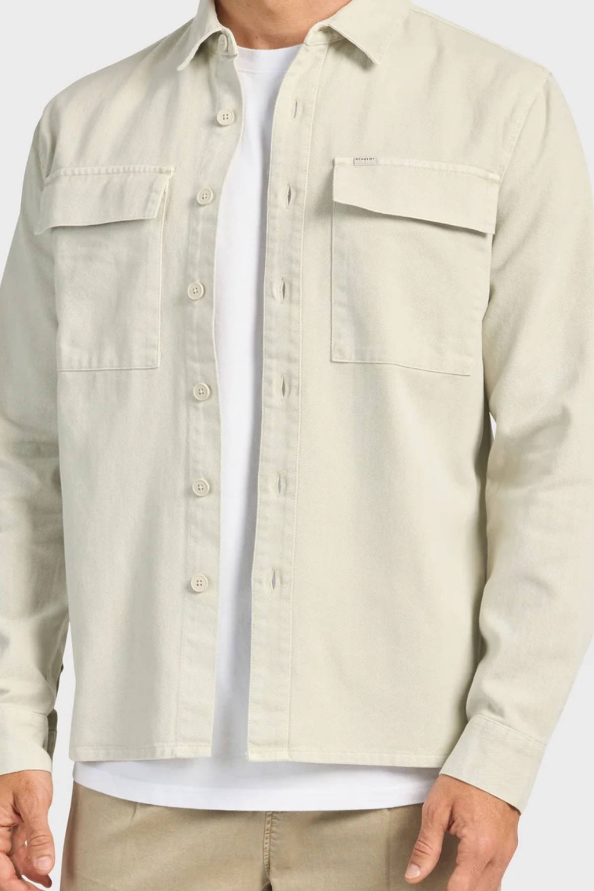 A man wears the Academy Brand Essential Overshirt in light beige cotton twill with double chest pockets, styled unbuttoned over a white tee and tan pants for a relaxed look.