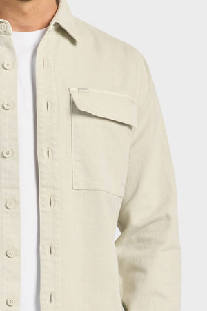 A person wears the Academy Brand Essential Overshirt in light beige cotton twill with a left chest flap pocket over a white t-shirt. Only the upper torso is visible against a plain background.