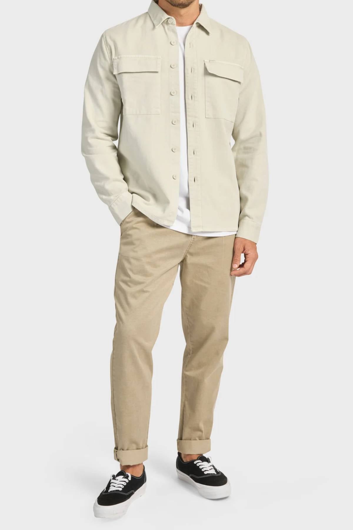 A man wearing the Academy Brand Essential Overshirt in light beige over a white T-shirt, paired with tan cuffed pants and black-and-white sneakers, stands against a plain white background. His head is not visible in the image.