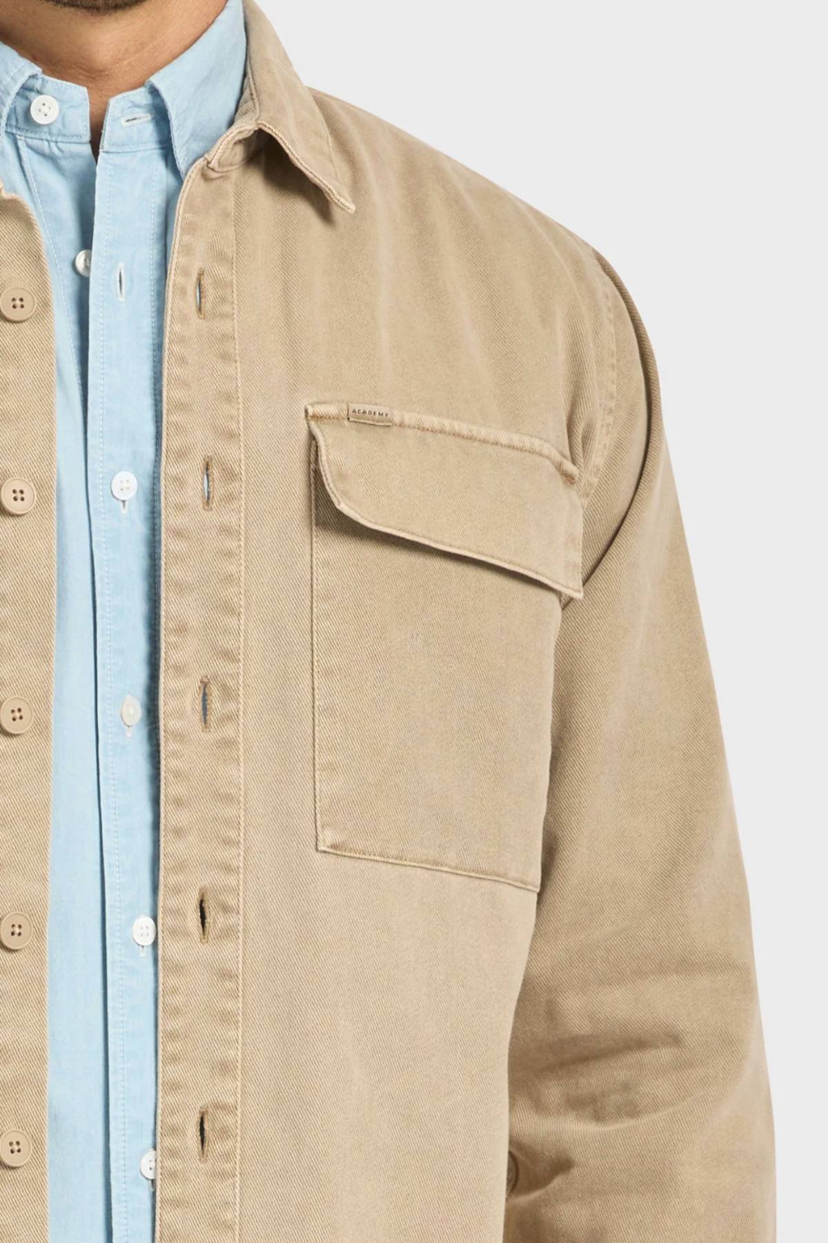 A person wears the Academy Brand Essential Overshirt in tan cotton twill with double chest pockets over a light blue button-up shirt, shown from shoulders to waist against a plain background.