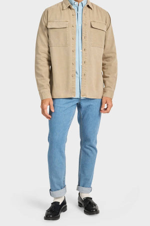 A person is shown from neck to feet wearing the Academy Brand Essential Overshirt in beige cotton twill with double chest pockets over a light blue collared shirt, light blue jeans, white socks, and black loafers.