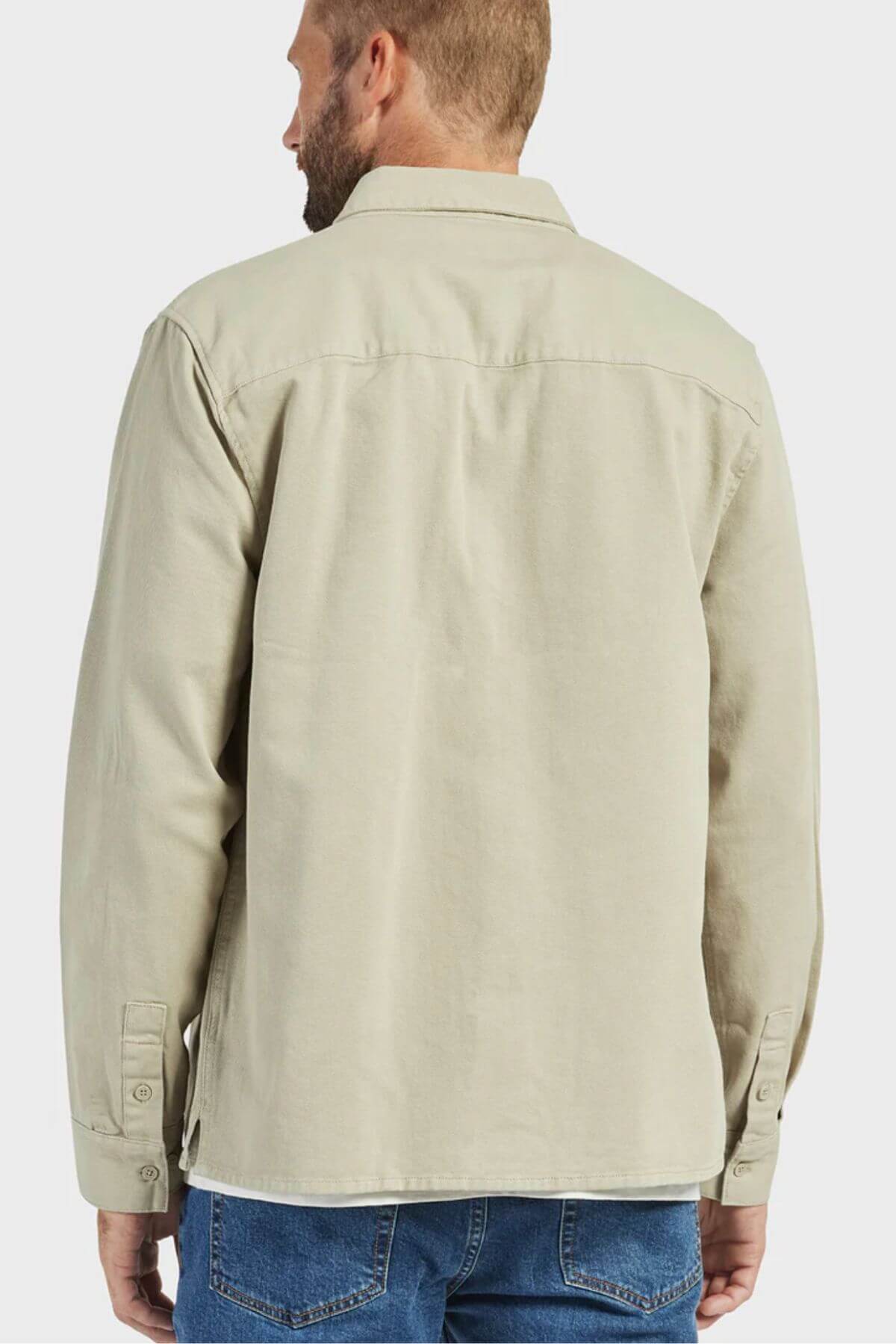 A person with short hair and a beard is standing facing away, wearing the Academy Brand Essential Overshirt in light beige, which features double chest pockets and buttoned cuffs, paired with blue jeans. The background is plain and light-colored.