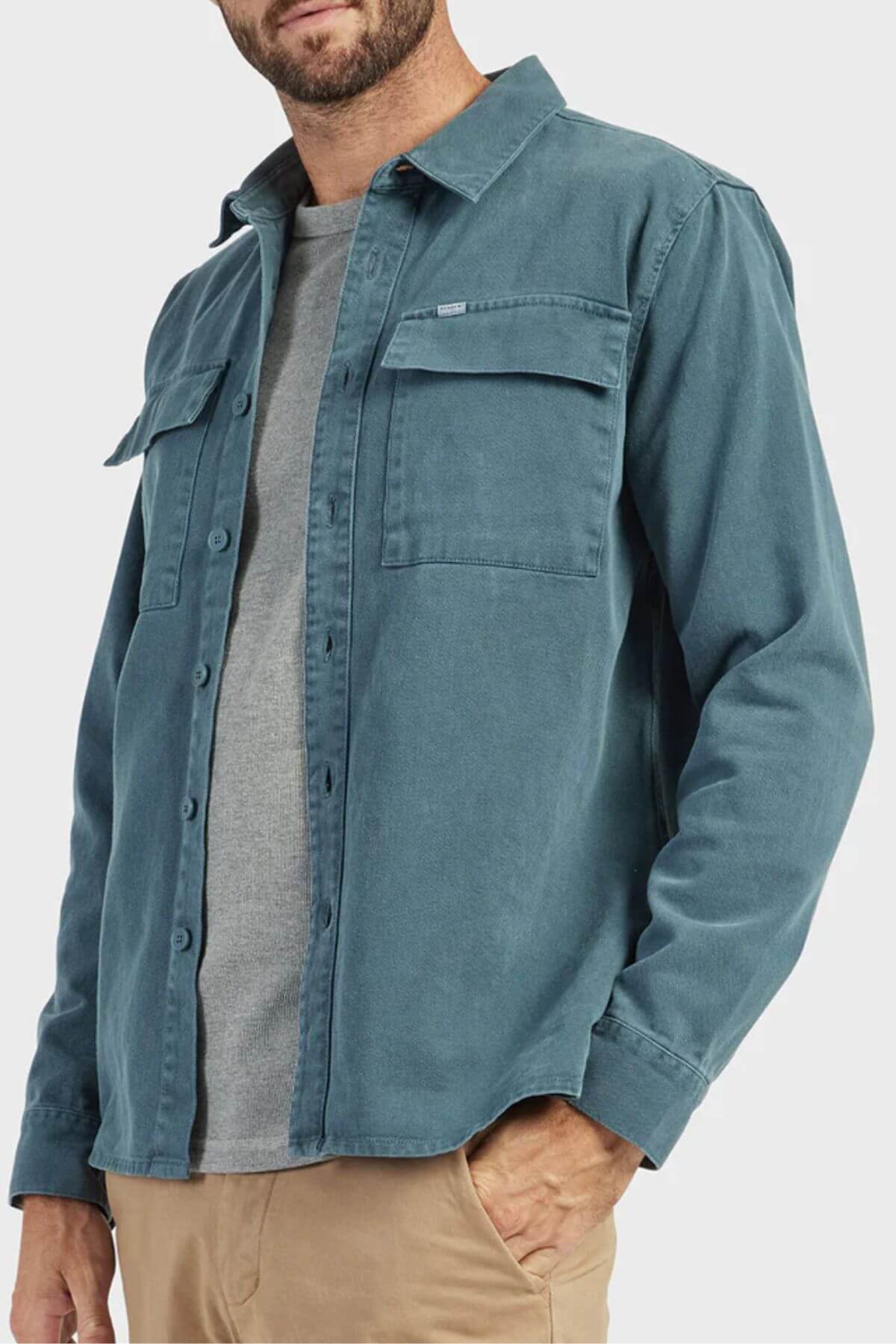 A man wears the Academy Brand Essential Overshirt in teal cotton twill with chest pockets over a gray t-shirt and beige pants, standing against a light background.