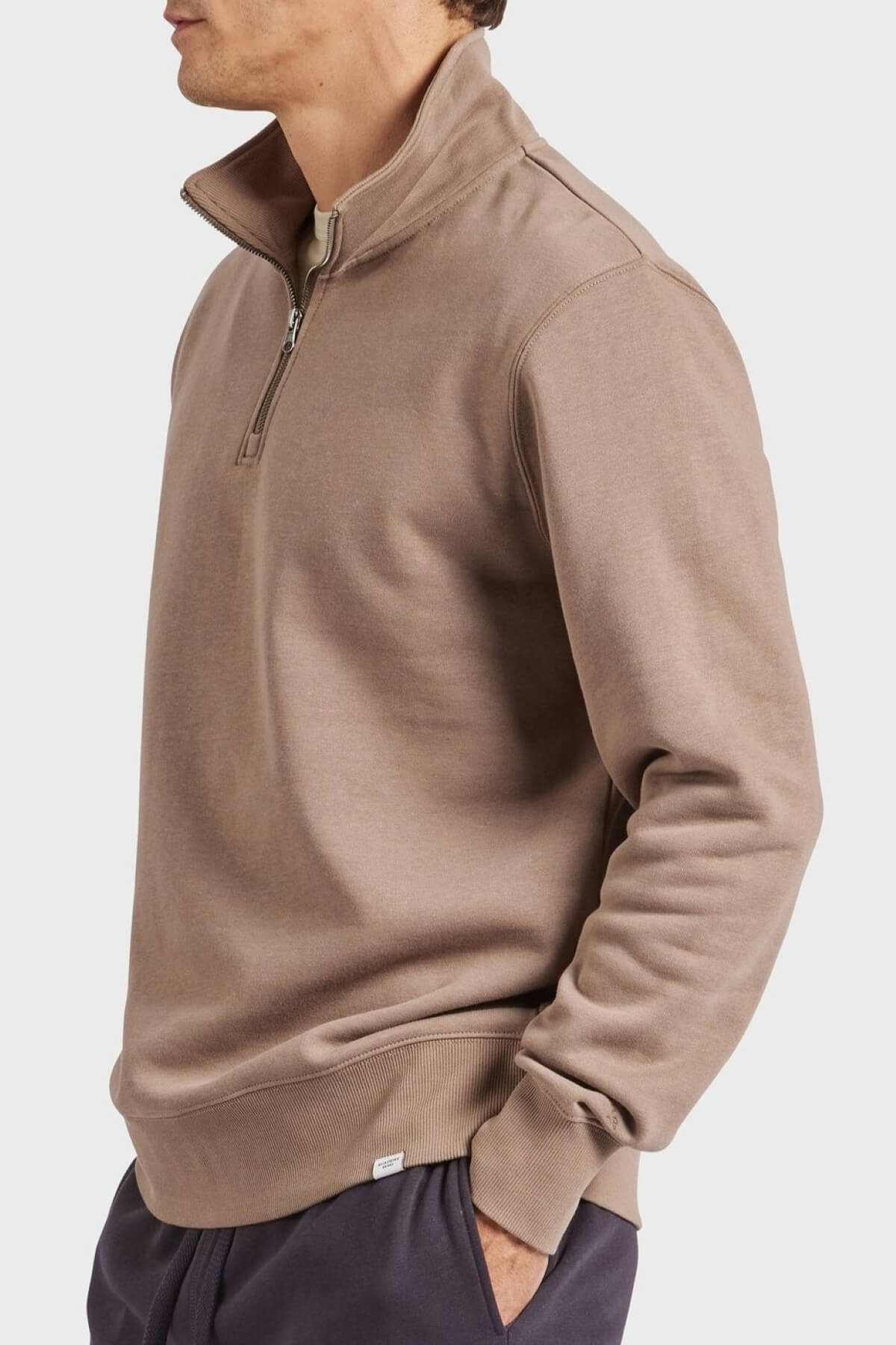 A person wearing the Academy Brand 1/2 Zip Sweat in taupe, with the zipper partially open, paired with dark pants. Their head is turned to the side, set against a plain white background.