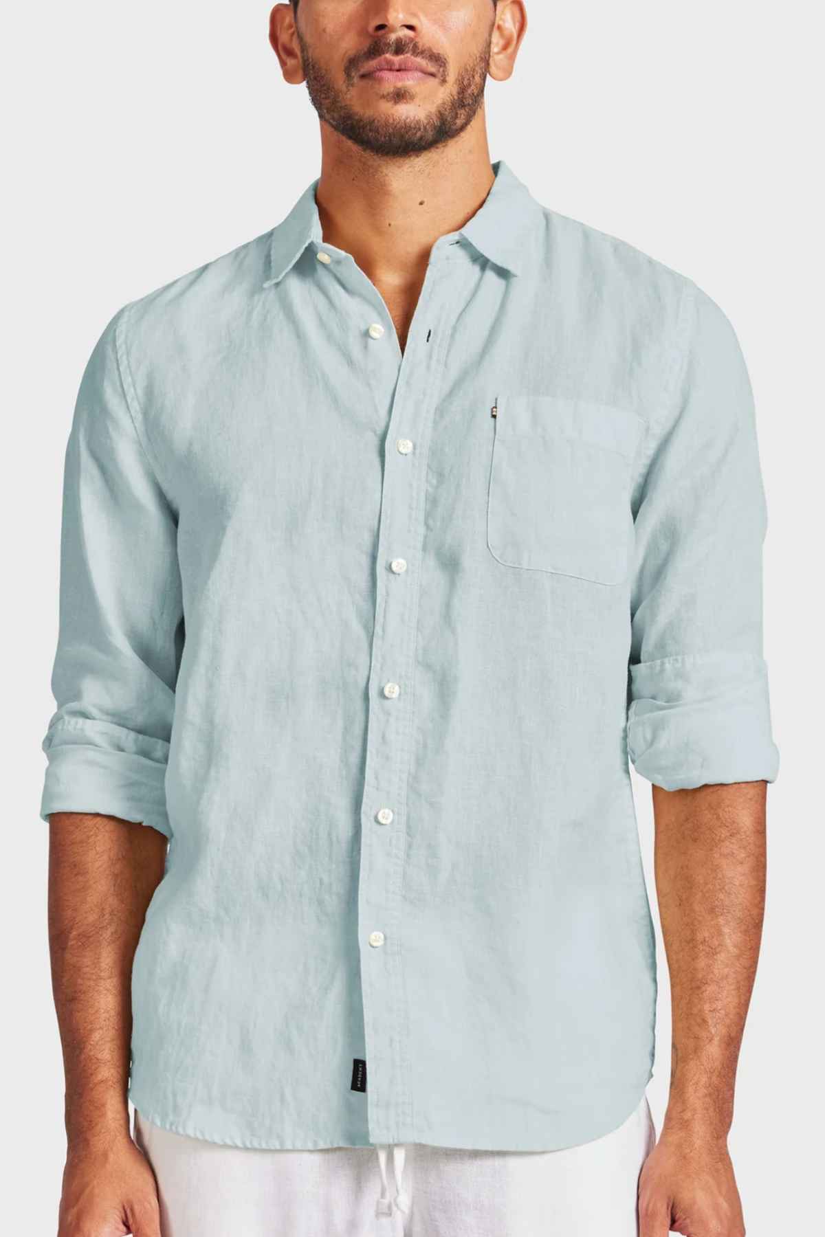 A man in an Academy Brand Hampton Linen Shirt, colored light blue, stands against a plain background, facing forward. Featuring rolled-up sleeves, a classic point collar, and a left-side pocket, the shirt adds refined touches to his look as he is visible from the waist up.