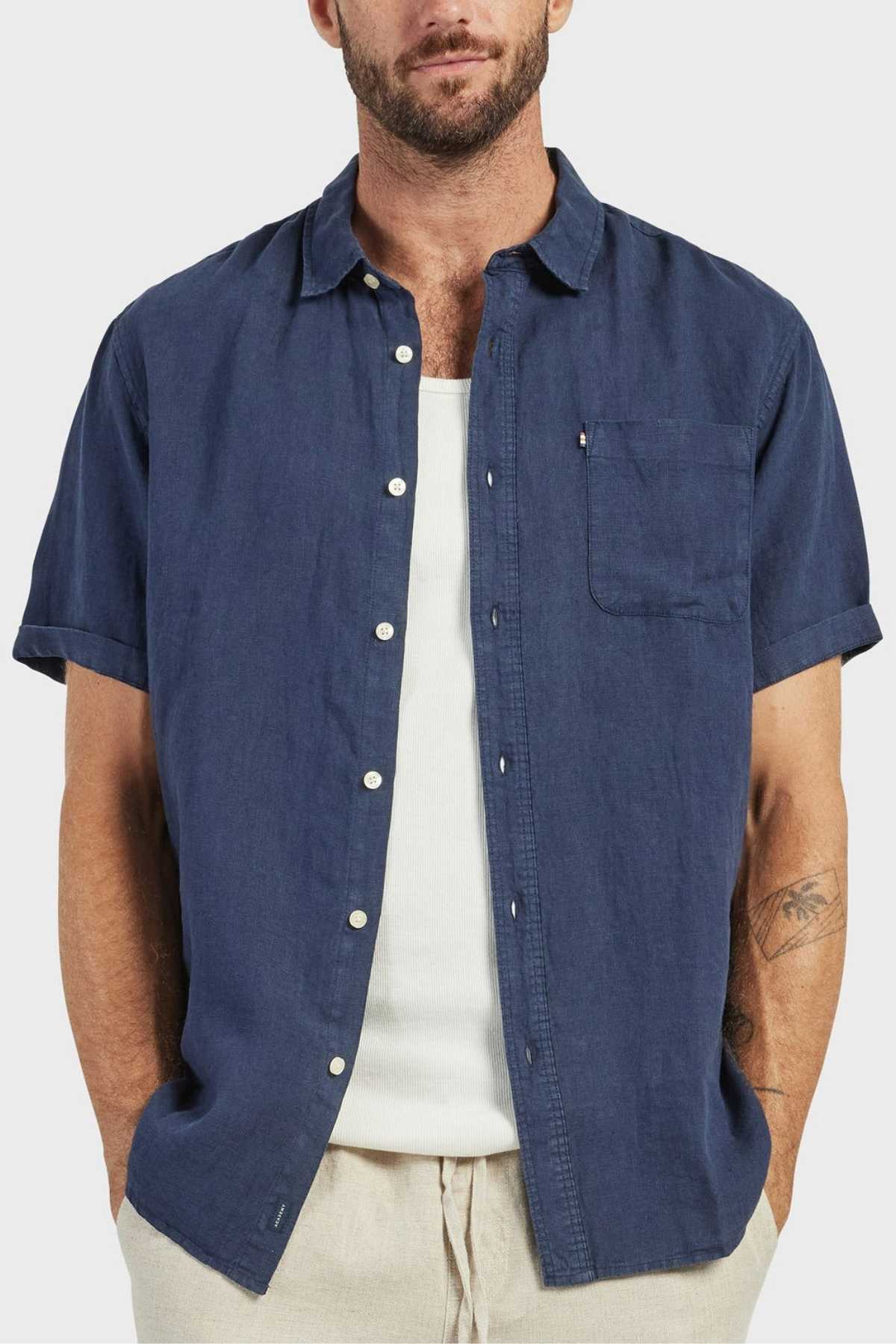 A man in the Academy Brand Hampton Linen Short Sleeve Shirt in navy, layered over a white tank top and beige pants, stands with hands in his pockets. His face is partly visible and he has a trimmed beard.