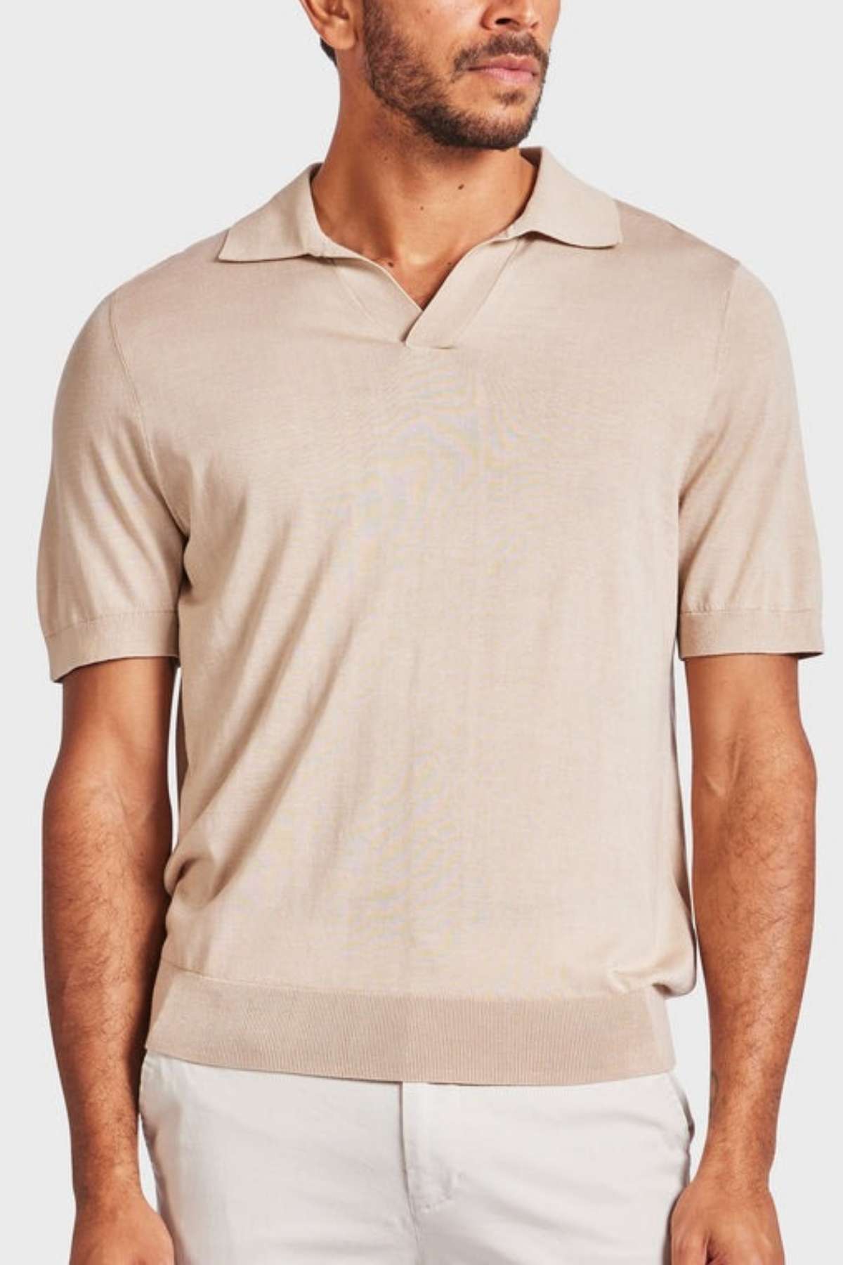 A man is sporting the vintage-inspired Academy Brand Irvine Knit Polo, featuring a beige short-sleeve design and a V-neck collar. The luxurious blend yarn of the shirt pairs seamlessly with light-colored pants, set against a plain white background.