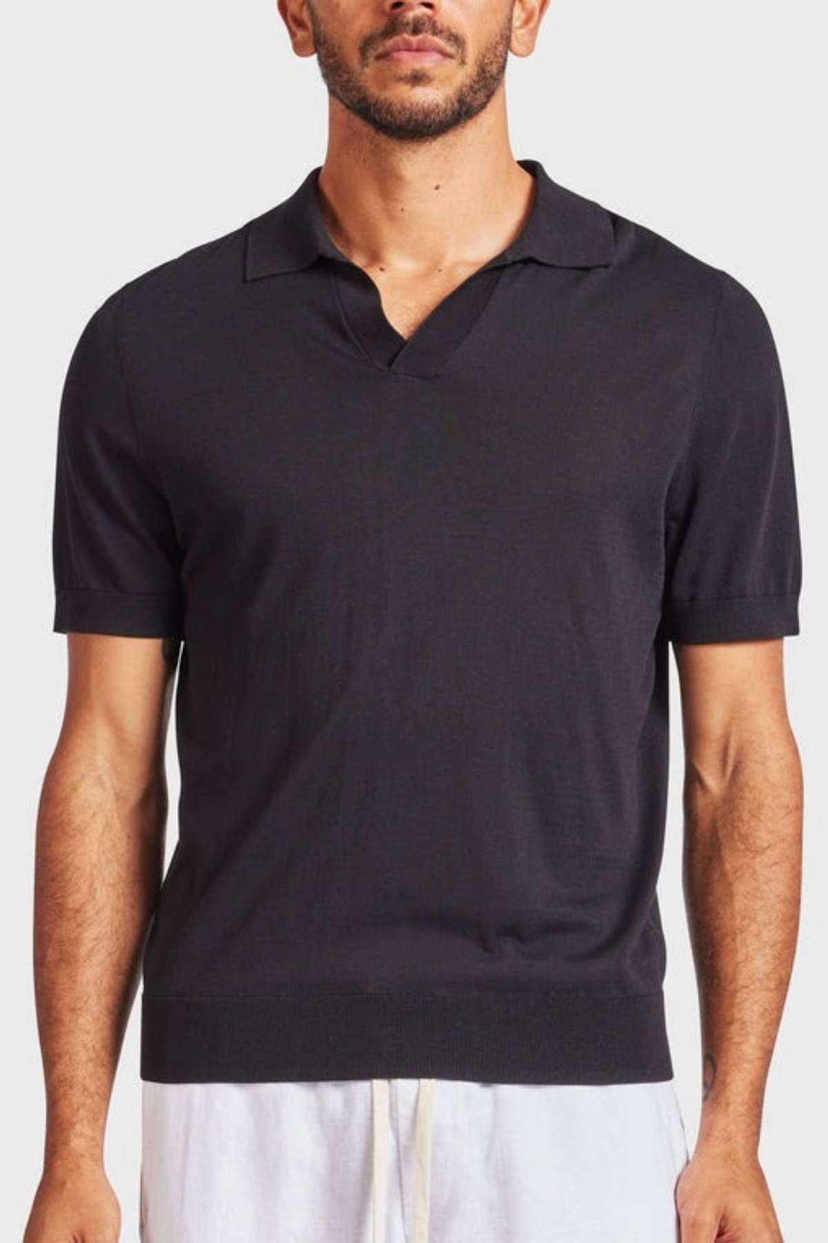A man with a short beard and mustache stands against a plain background, modeling the Academy Brand Irvine Knit Polo, which features a V-neckline and short sleeves. He pairs the vintage-inspired polo with white pants, his arms slightly bent at the elbows.