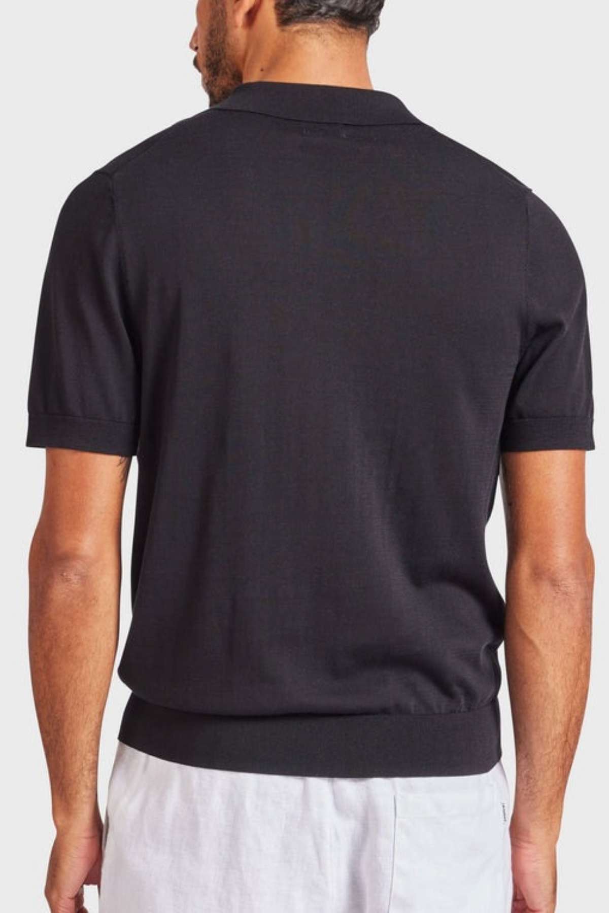 A man with short hair is turned away from the camera, sporting an Academy Brand Irvine Knit Polo—a vintage-inspired black short-sleeved top—and light-colored pants. The backdrop is a plain, light gray tone.