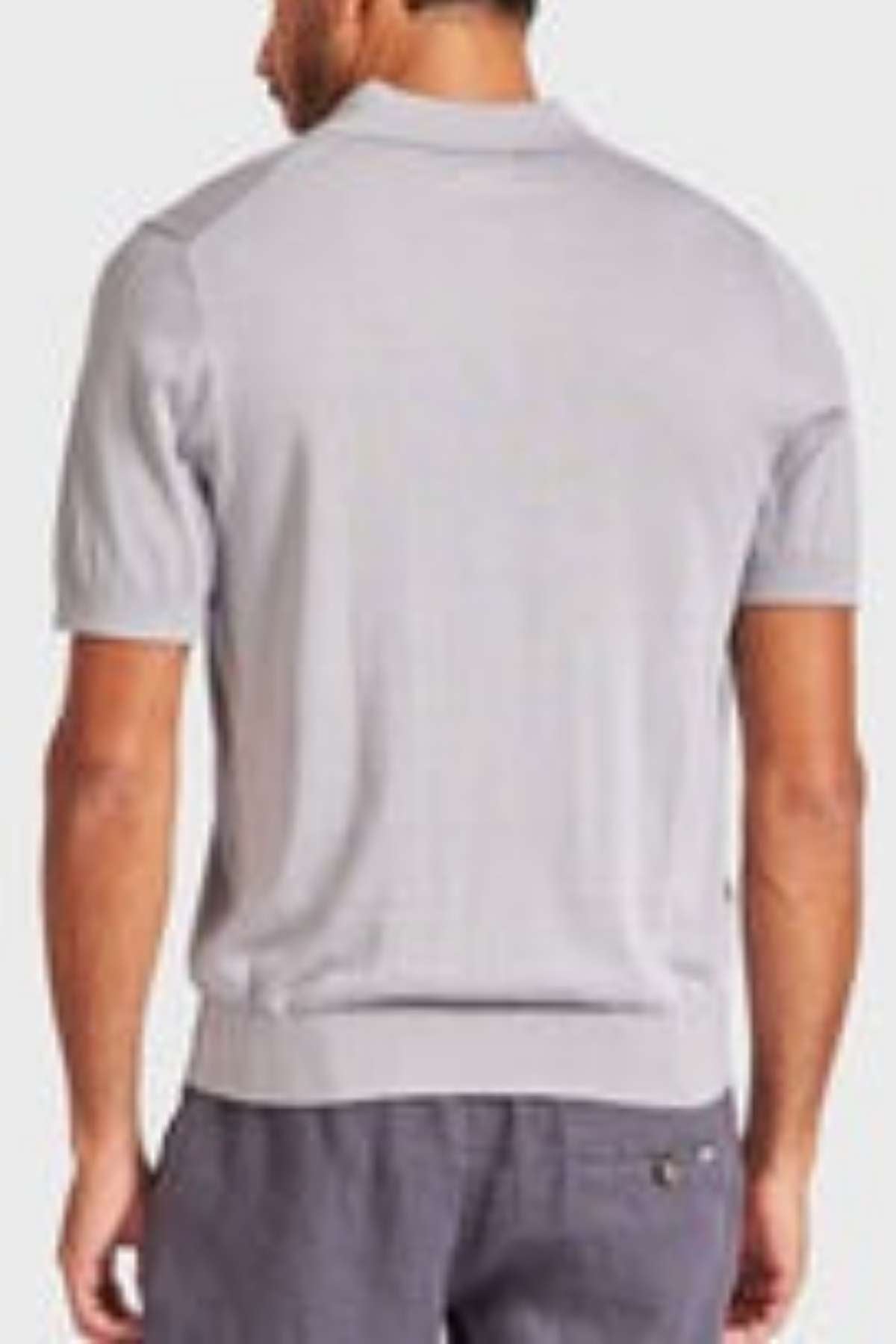 A man facing away from the camera is wearing a light gray Academy Brand Irvine Knit Polo and darker gray pants. The background is plain and unobstructed.