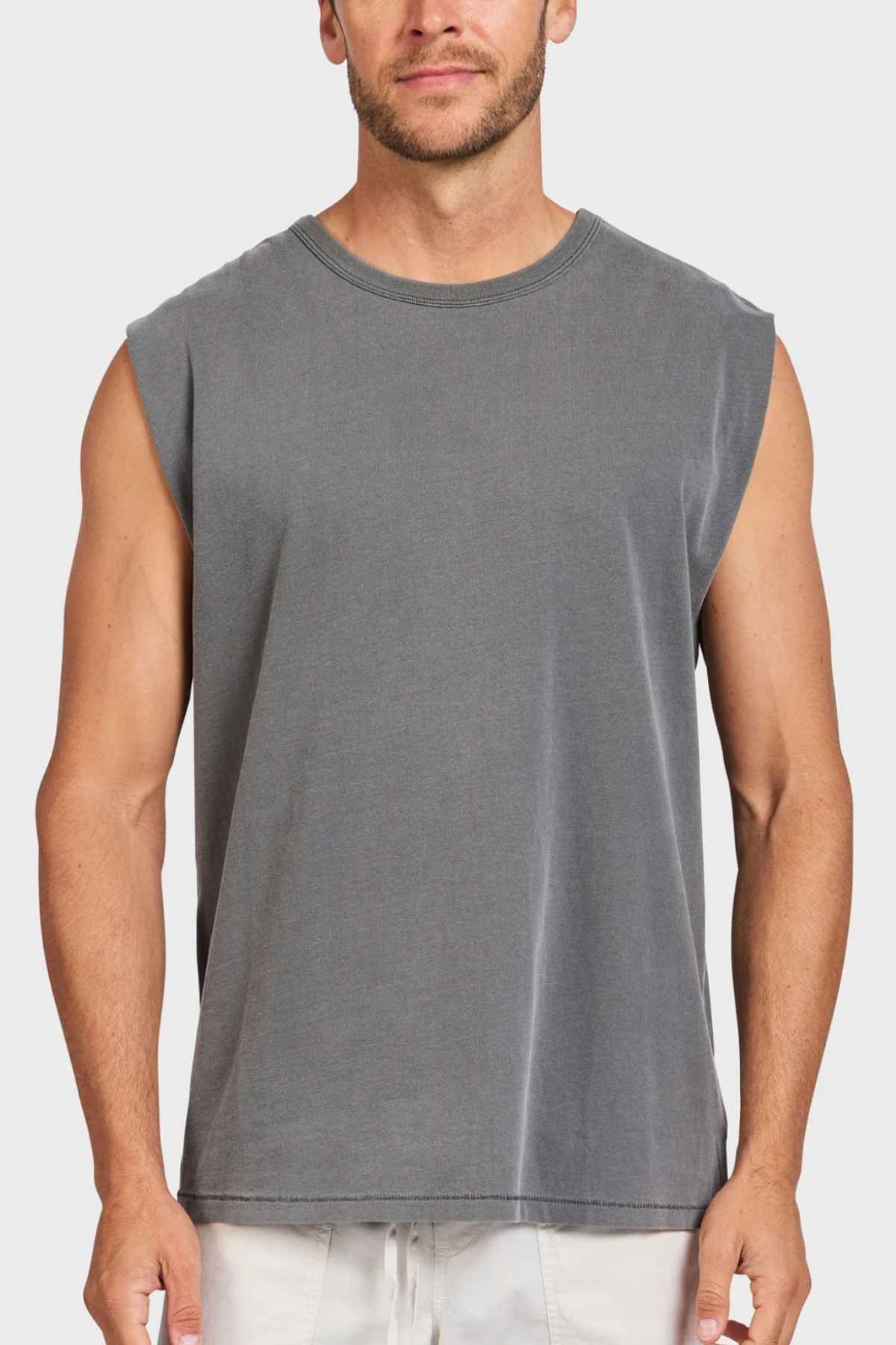 A man stands against a white background, dressed in the Academy Brand Jimmy Muscle Tee in gray along with light-colored shorts. With short hair and a light beard, his arms rest relaxed at his sides while he gazes forward. The tee, crafted from 100% cotton, offers both comfort and style.
