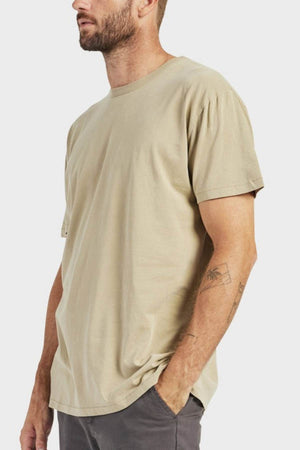 A man with a short beard wears a loose fit Academy Brand Jimmy Tee made from 100% cotton and dark gray pants. His left hand, showing forearm tattoos, is in his pocket. The background is plain white.