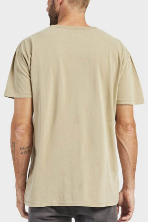 A person with short hair and left forearm tattoos is seen from behind wearing the Academy Brand Jimmy Tee, a plain beige, loose-fit 100% cotton t-shirt, paired with gray pants against a neutral background.