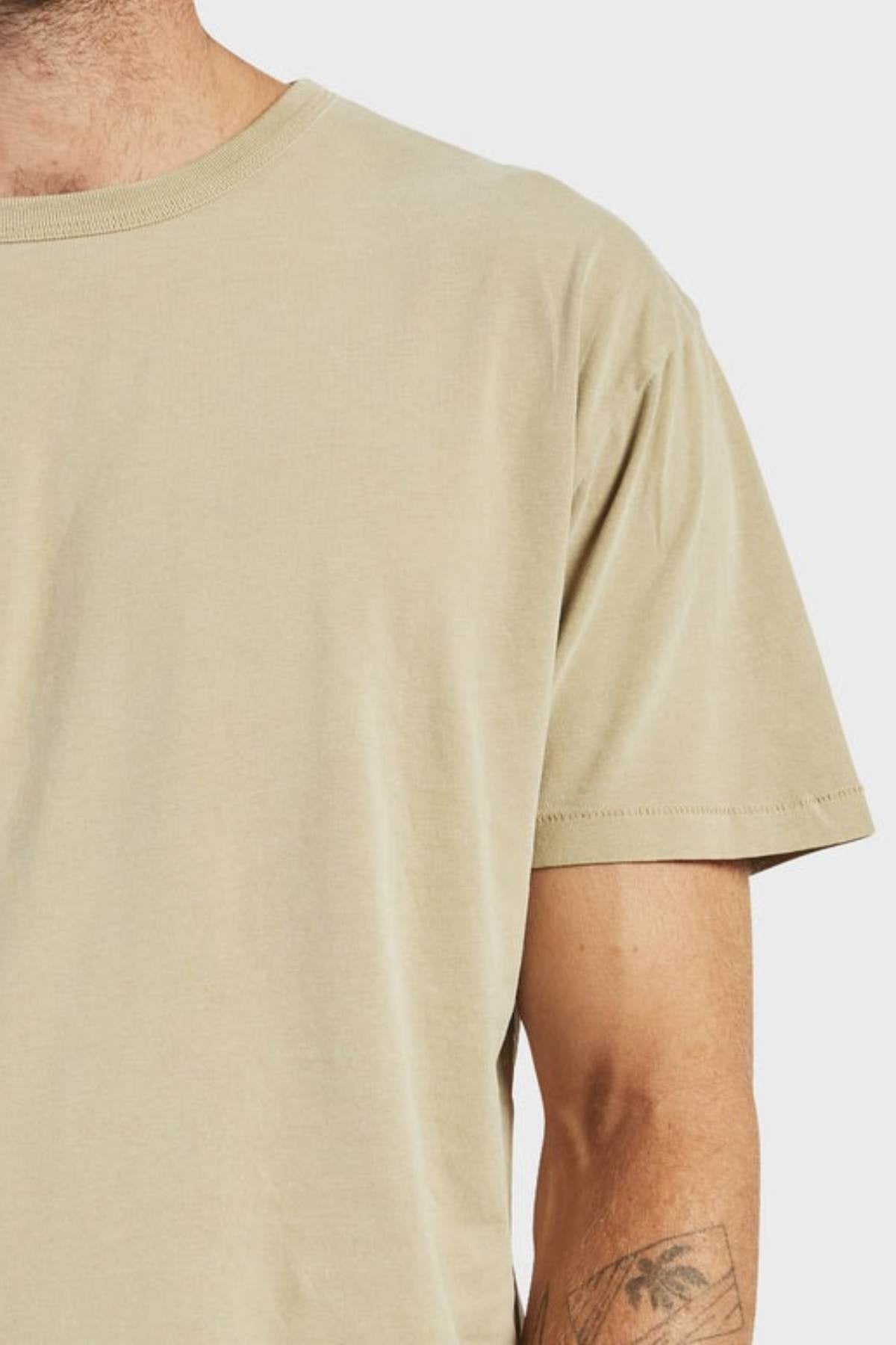 A person wears an Academy Brand Jimmy Tee, a loose-fitting, 100% cotton plain beige t-shirt. Only their lower face, upper torso, and tattooed left arm are seen against a light background.