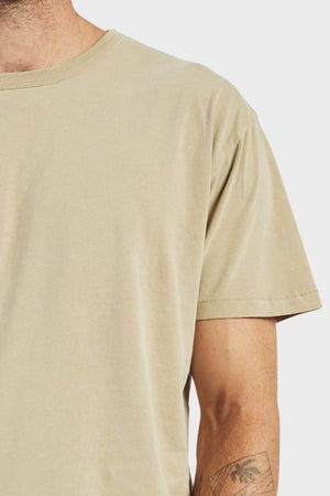 A person wears an Academy Brand Jimmy Tee, a loose-fitting, 100% cotton plain beige t-shirt. Only their lower face, upper torso, and tattooed left arm are seen against a light background.