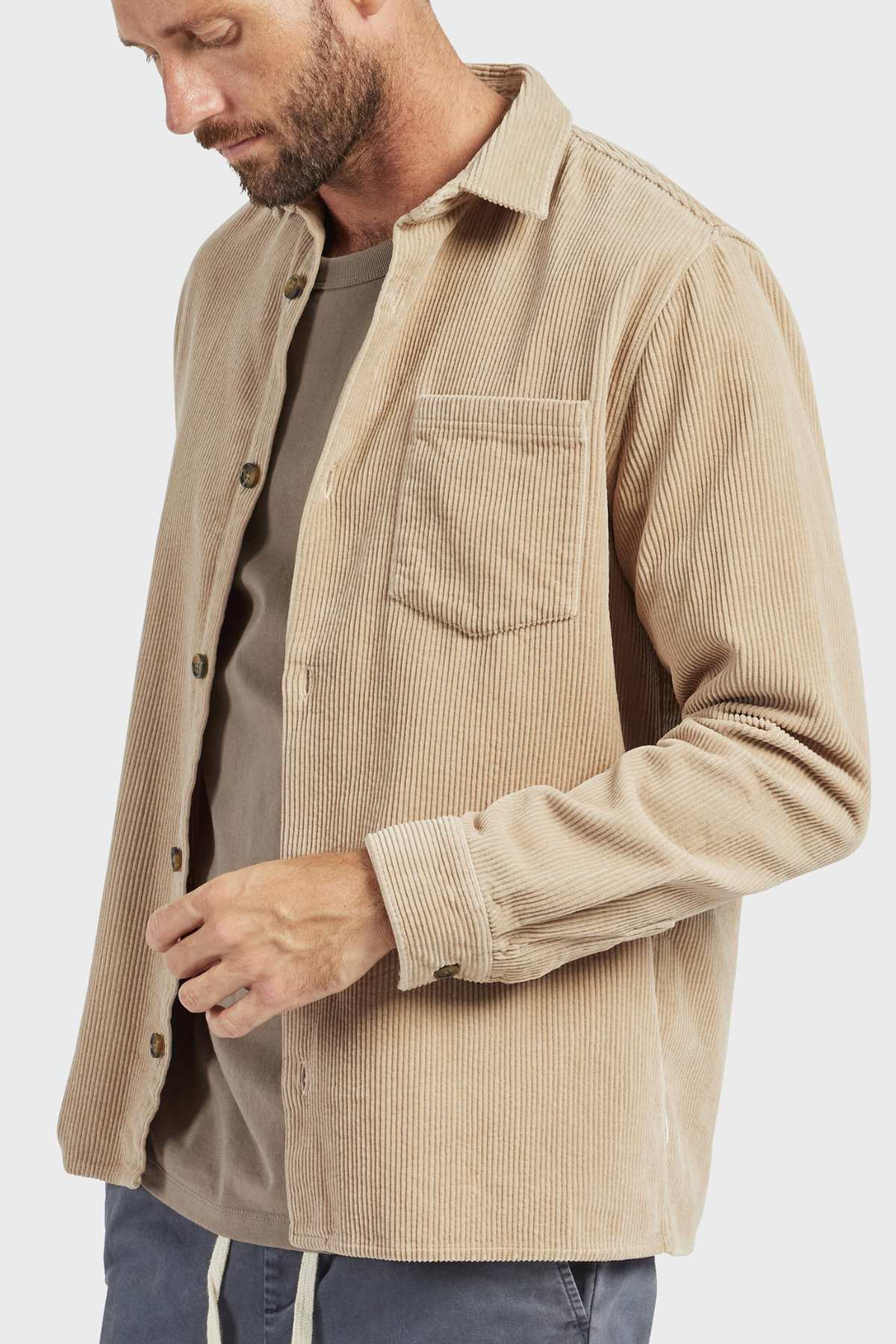 A bearded man wearing the stylish Academy Brand Lebowski Cord Overshirt, made of mid-weight 100% cotton corduroy, over a gray t-shirt looks down while adjusting his sleeve. The plain white background highlights his attire.