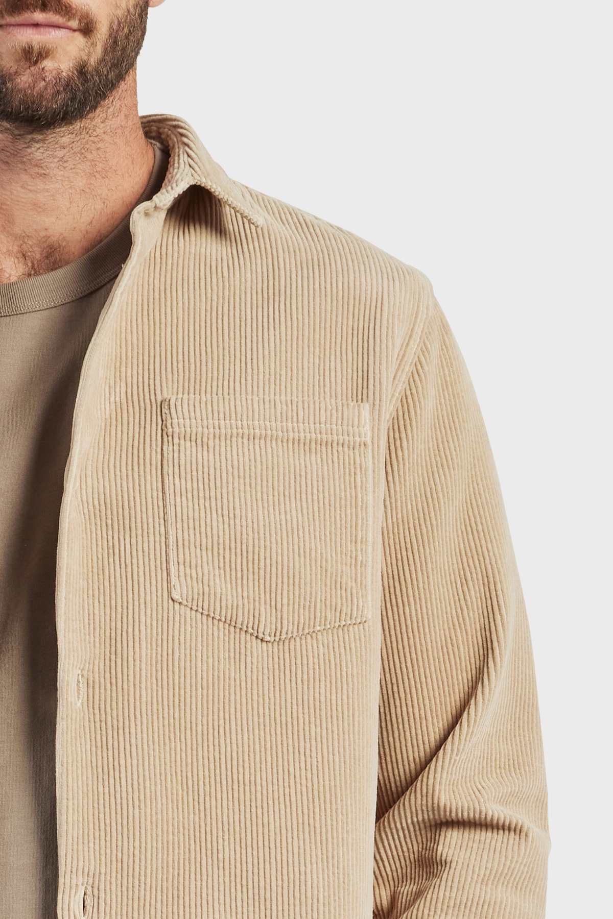 A close-up of a person wearing the Academy Brand Lebowski Cord Overshirt, a stylish, mid-weight shirt made of 100% cotton, over a taupe-colored T-shirt. The overshirt features a chest pocket and is slightly unbuttoned. The image captures only the upper torso and part of the person's face against a neutral background.