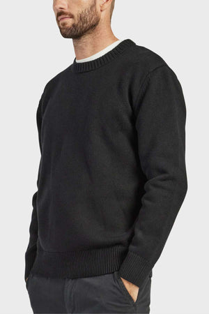 A bearded man is wearing an Academy Brand Malibu Crew sweater in black over a white undershirt. With his hands tucked into the pockets of his dark trousers, he poses against a plain white background, exemplifying a cozy and stylish aesthetic.