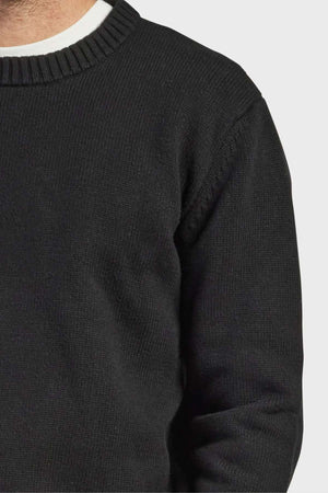 A person, shown from the shoulders to the mid-torso, is wearing the high-quality Academy Brand Malibu Crew sweater in black, with a faintly visible white undershirt at the collar. The cozy and stylish look is highlighted against a plain, light-colored background.