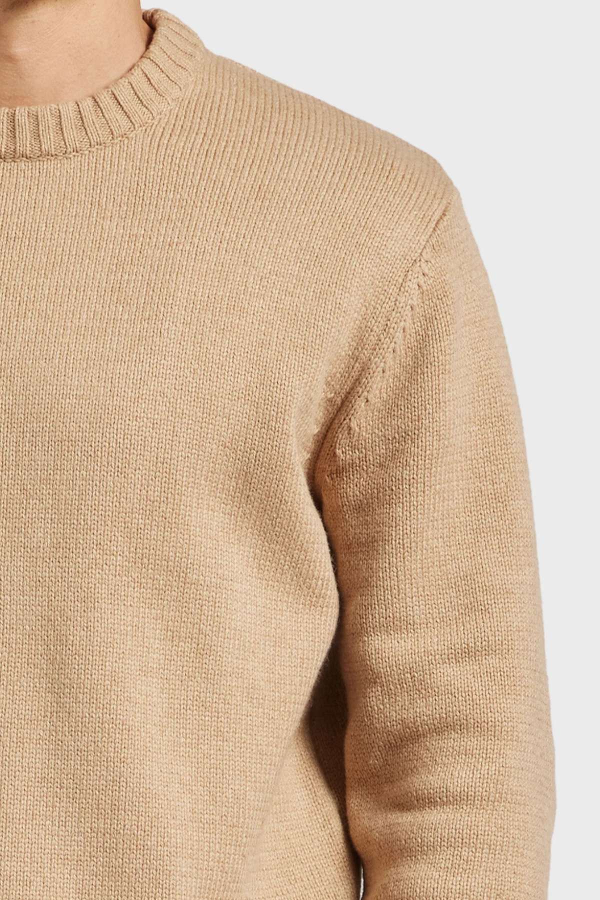 A close-up of a person wearing an Academy Brand Malibu Crew light beige knitted sweater, highlighting the slightly oversized fit. The focus is on the torso and part of the left arm. The sweater features a ribbed neckline and appears cozy and soft against a plain white background.