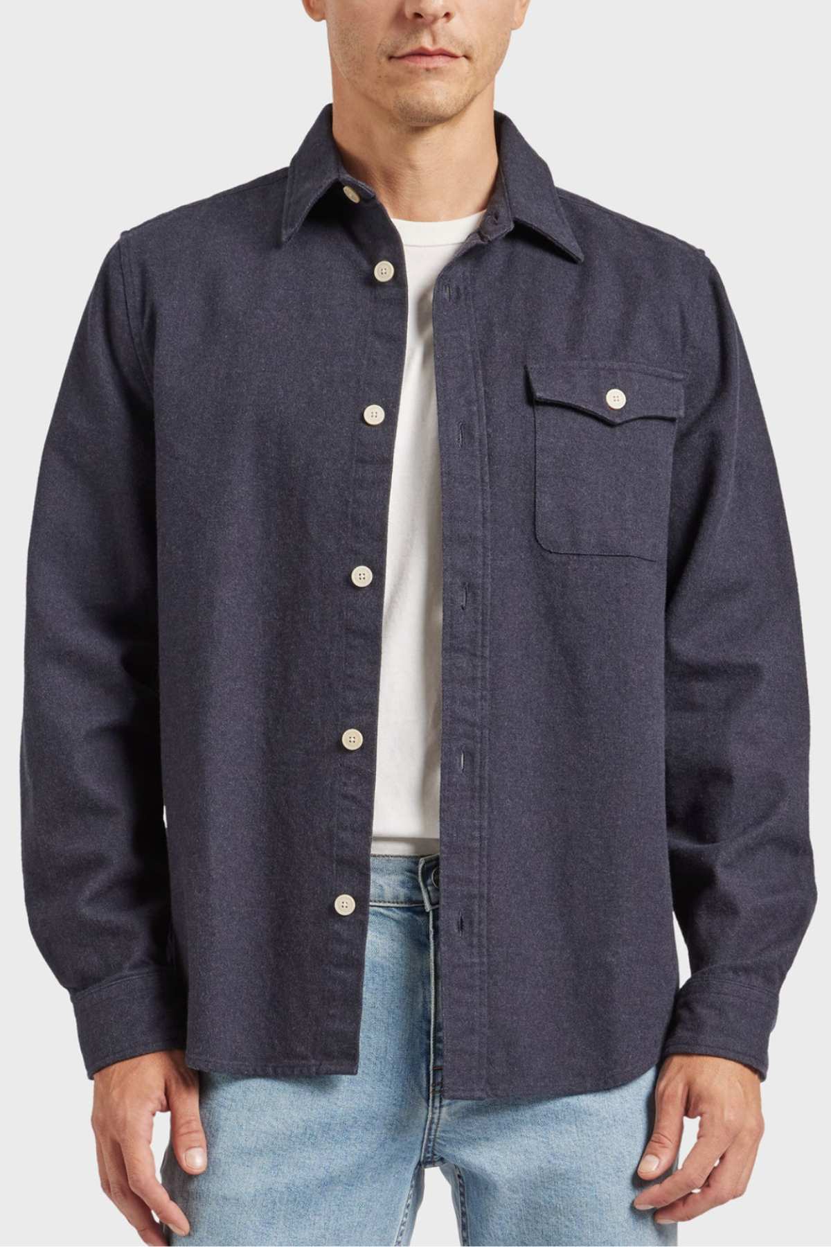 Clad in an Academy Brand Oakland Overshirt in dark grey, made from premium yarn-dyed twill and 100% cotton, a person showcases its design which includes a chest pocket on the left side with a button closure. The stylish look is complemented with light blue jeans and a white t-shirt underneath, making the ensemble pop against the plain white background.