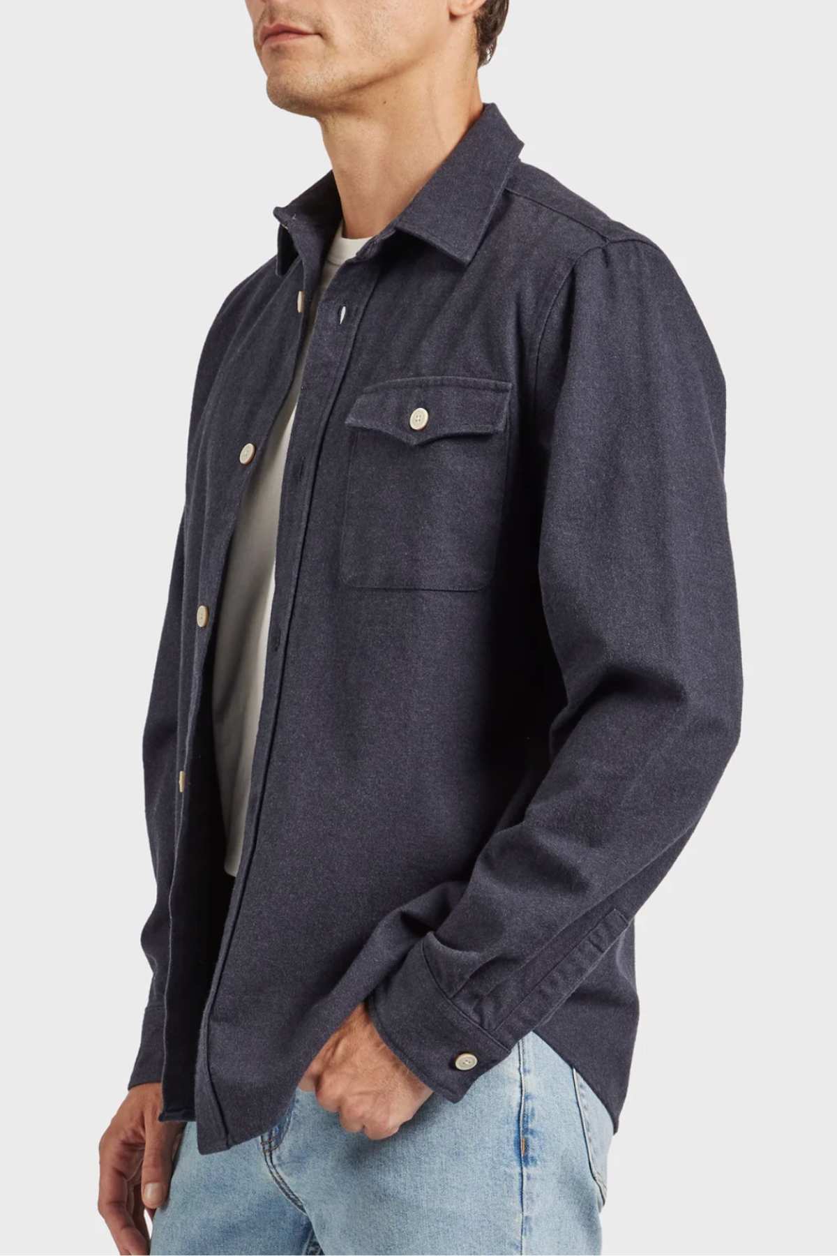 A person stands against a plain white background, wearing the Academy Brand Oakland Overshirt crafted from premium yarn dye twill over a light gray T-shirt and light blue jeans. Their head is slightly turned to the right, and their left hand rests inside the pocket of their jeans.