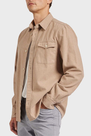 A person with light skin and short hair is wearing a tan Academy Brand Oakland Overshirt, which is crafted from premium yarn dye twill and features a chest pocket. They are also in light gray pants, standing with their right hand in their pants pocket, facing slightly to the right against a plain white background.