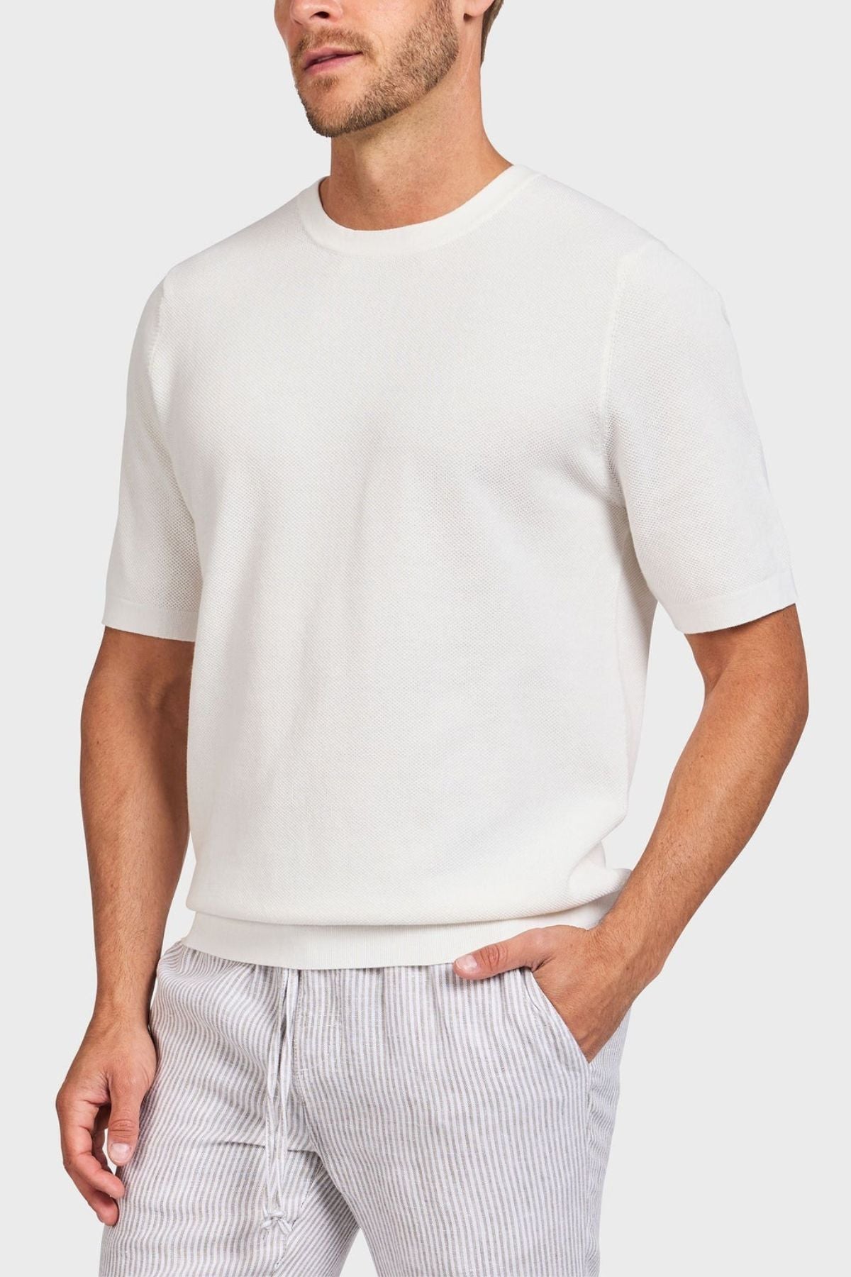 Wearing a simple white Academy Brand Riley Knit Tee, recognized for its lightweight breathable fabric, and light gray striped pants, an individual stands with a hand in their pocket against a neutral backdrop.
