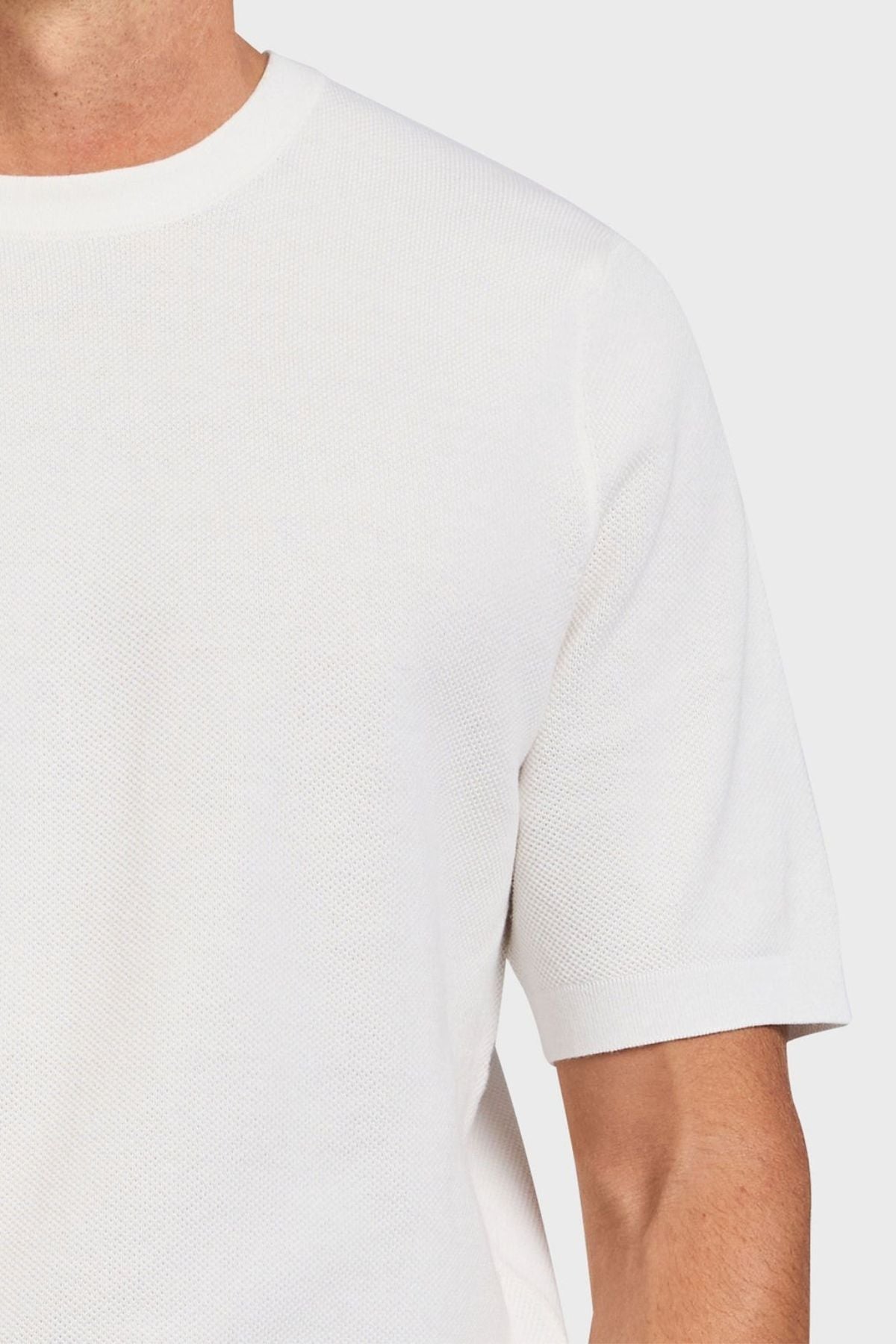 A person wears an Academy Brand Riley Knit Tee in plain white with short sleeves. The light gray background highlights the premium textured stitch of its lightweight, breathable fabric around the neckline and upper part.