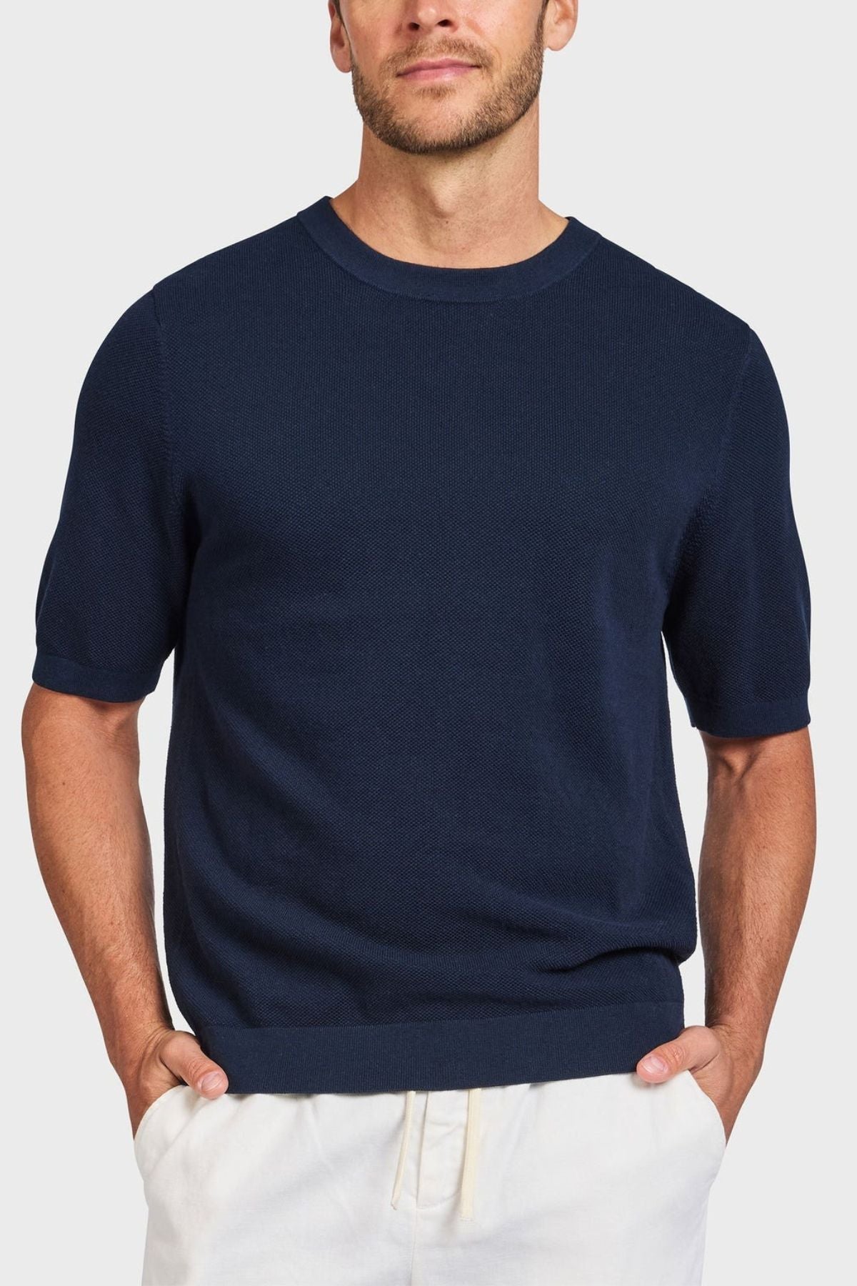 A man in a dark blue Academy Brand Riley Knit Tee and light pants stands with hands in pockets against a white background, highlighting the premium textured stitch that adds elegance and style.