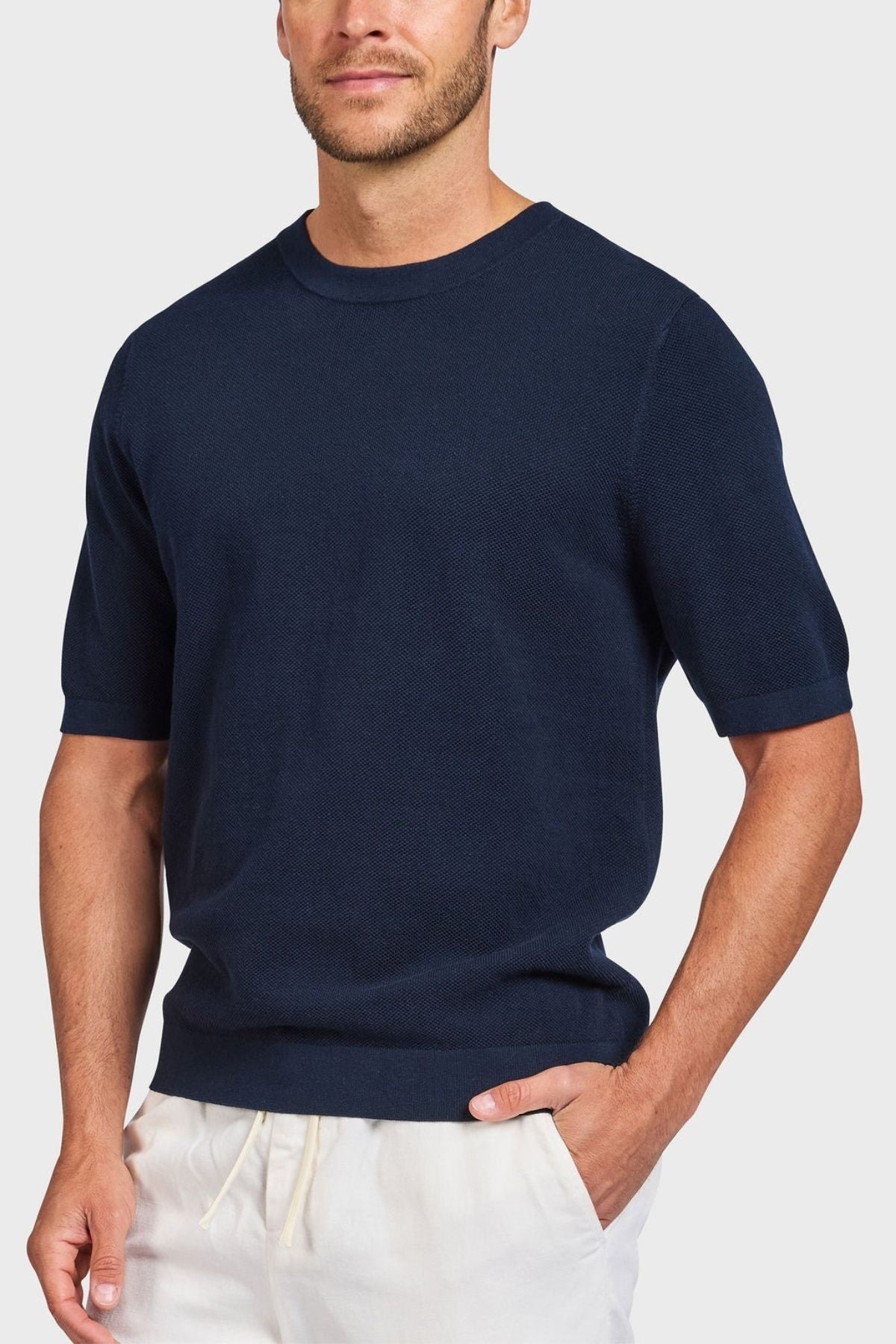 A man in a navy Academy Brand Riley Knit Tee, made from breathable fabric, and light beige pants stands against a plain, light background, casually posing with one hand on his hip.