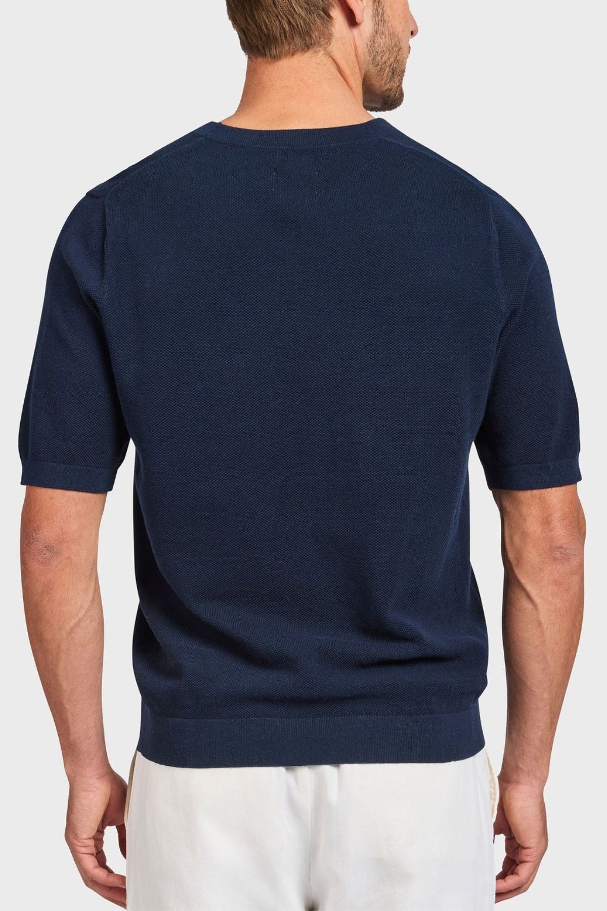 A man faces away from the camera, highlighting an Academy Brand Riley Knit Tee in navy blue made from breathable fabric, paired with crisp white pants against a plain light gray background.