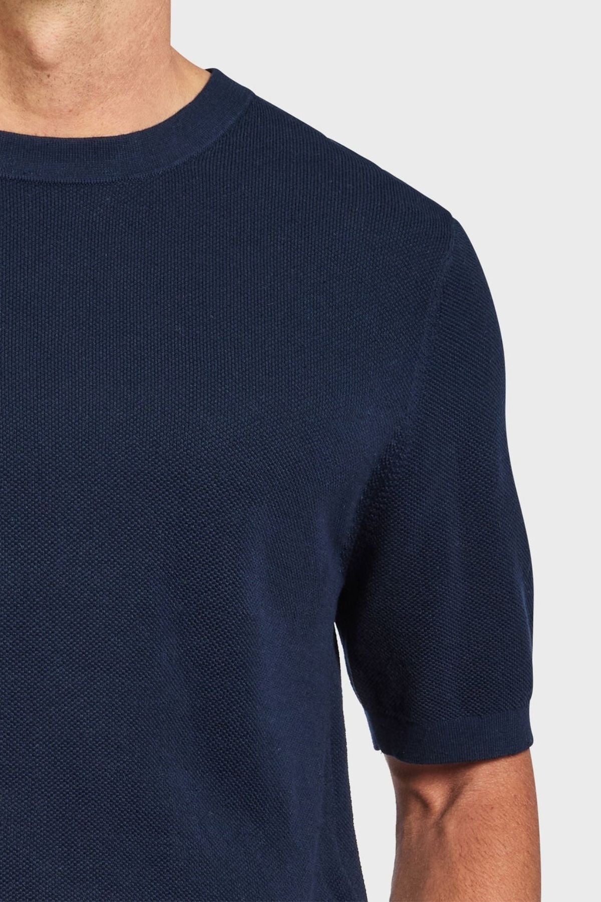 A person wearing a dark blue Academy Brand Riley Knit Tee is shown from the neck to the torso, with their left arm slightly visible. The textured stitch complements its breathable fabric against a plain, light-colored background.