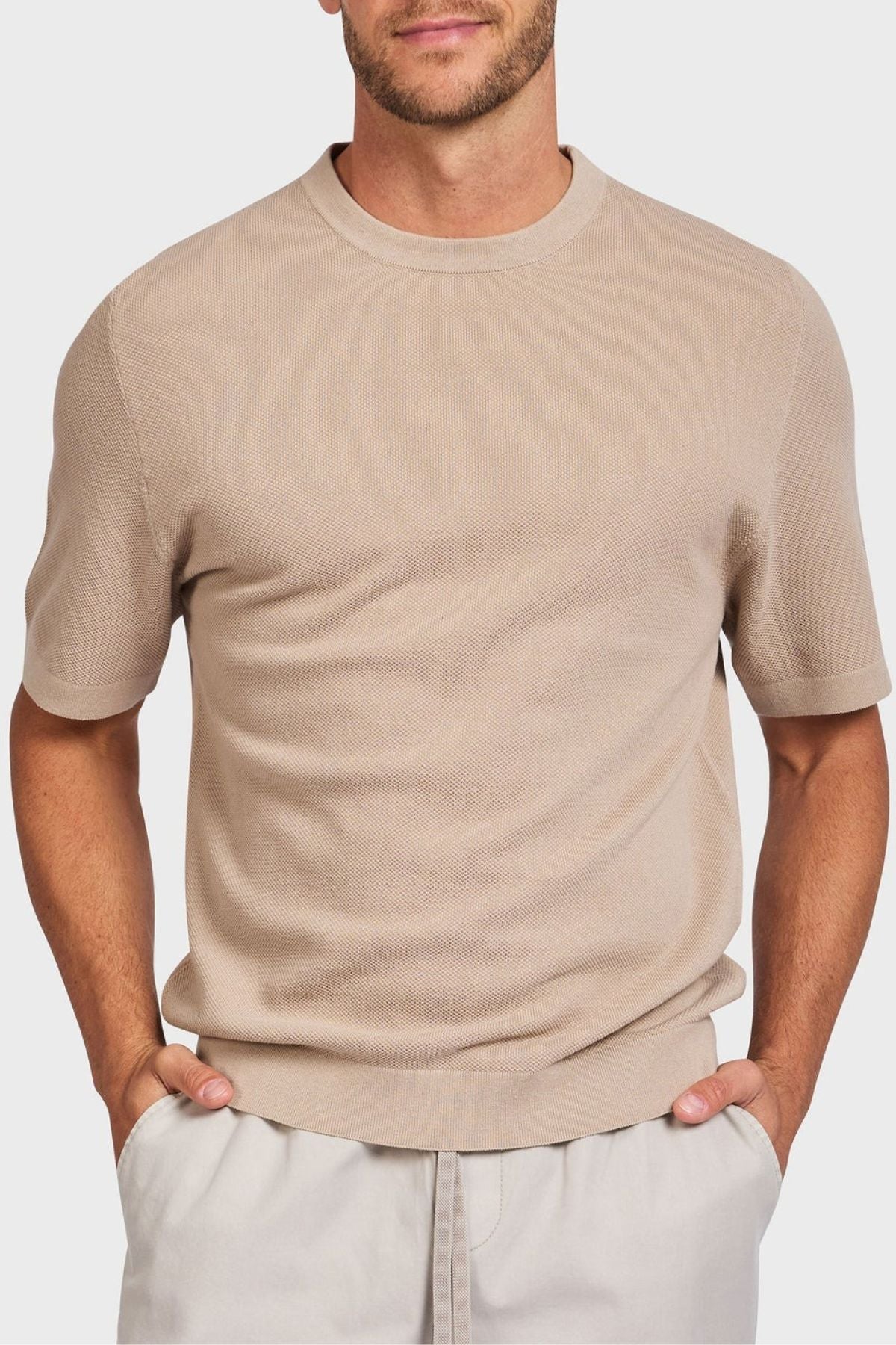 A person stands against a plain background, hands in pockets, wearing the Academy Brand Riley Knit Tee, a beige short-sleeve sweater made from lightweight breathable fabric, paired with light gray pants.
