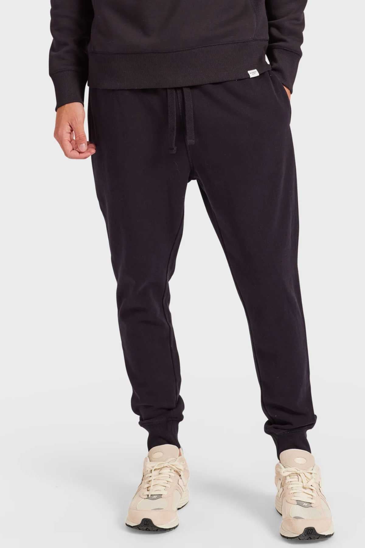 Wearing Academy Brand Sweat Pants featuring an elastic waist, someone pairs them with cream sneakers. Their hand is in their pocket, highlighting comfort and style against a plain white background.