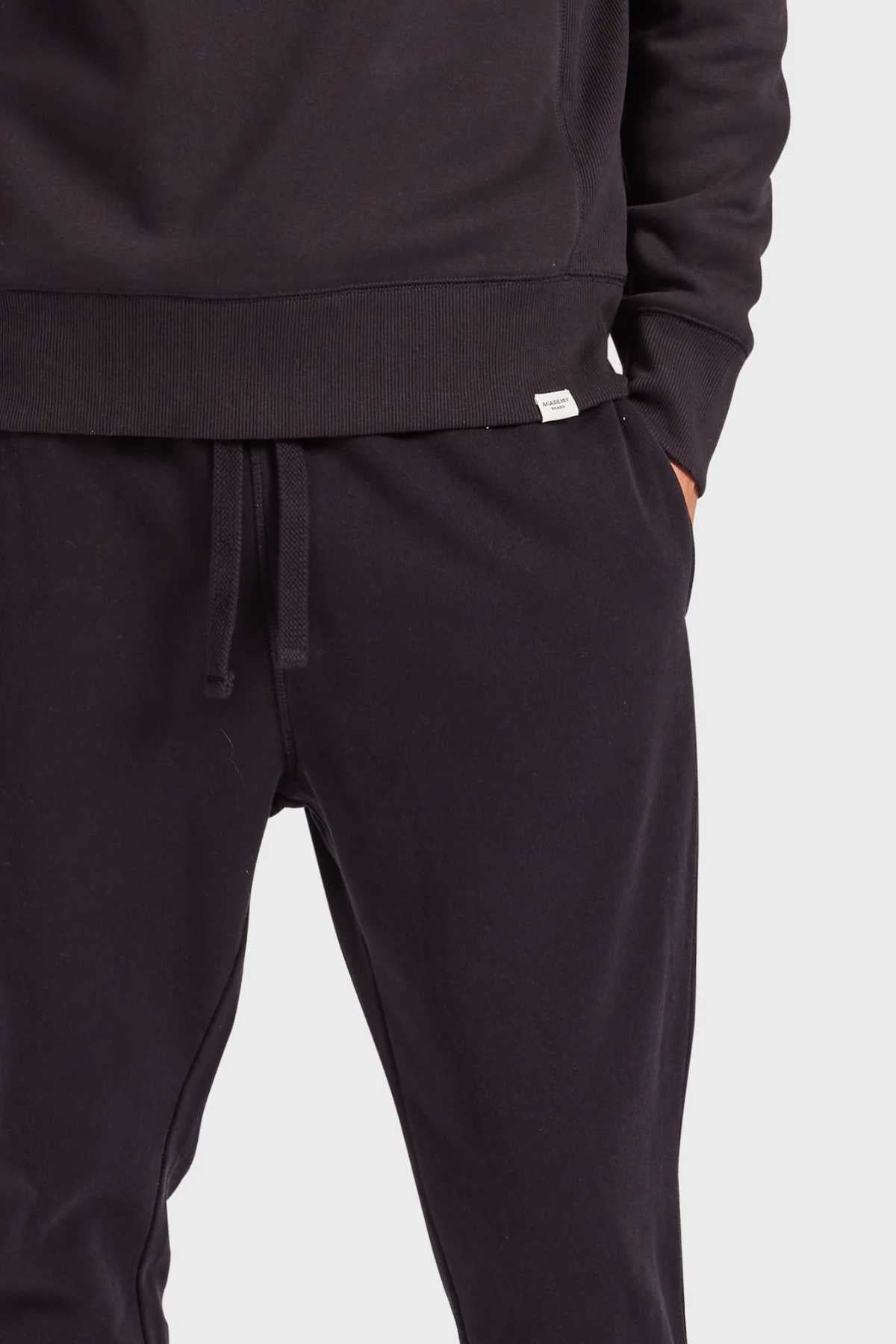 Wearing a black hoodie and Academy Brand Sweat Pant featuring an elastic waist and premium brushed fleece, a person slips their hands into the pockets. Only their torso and upper legs are visible against a plain white background.