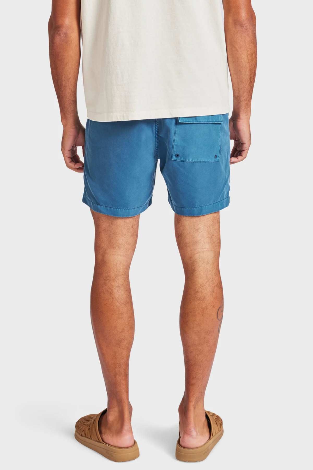 A person stands facing away, wearing a cream T-shirt, Academy Brand Teamster Boardy vintage-look blue quick dry swim shorts with a back pocket, and tan sandals against a plain light background.