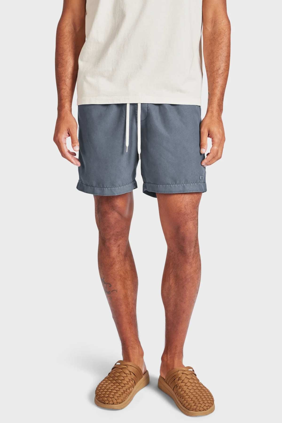 A person stands against a plain white background wearing the Academy Brand Teamster Boardy gray tailored fit quick dry swim shorts made from recycled polyester, paired with a cream t-shirt and brown woven slip-on shoes. The image shows them from shoulders to feet.