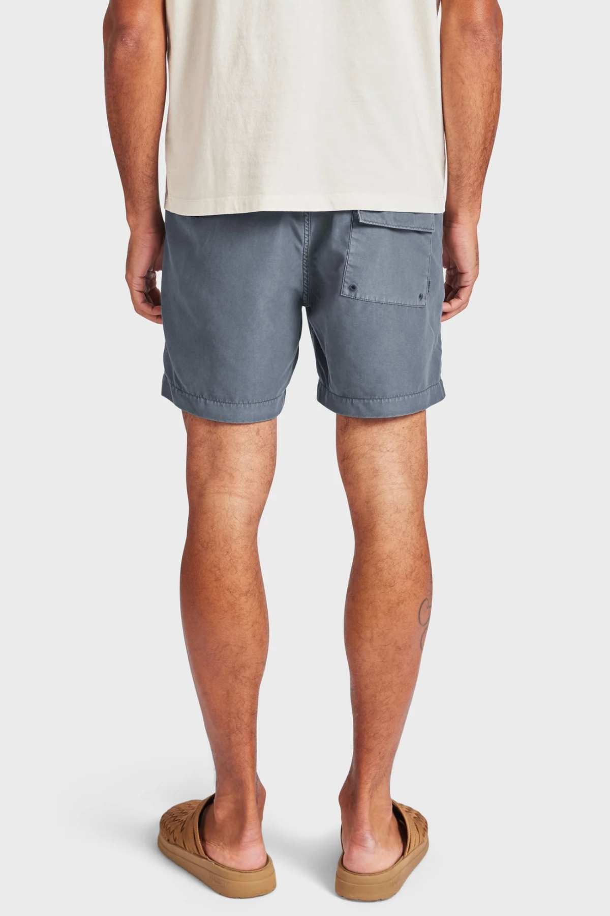 A person stands facing away, wearing the Academy Brand Teamster Boardy tailored fit blue-gray quick dry swim shorts with a back pocket, beige t-shirt, and tan sandals against a plain white background.