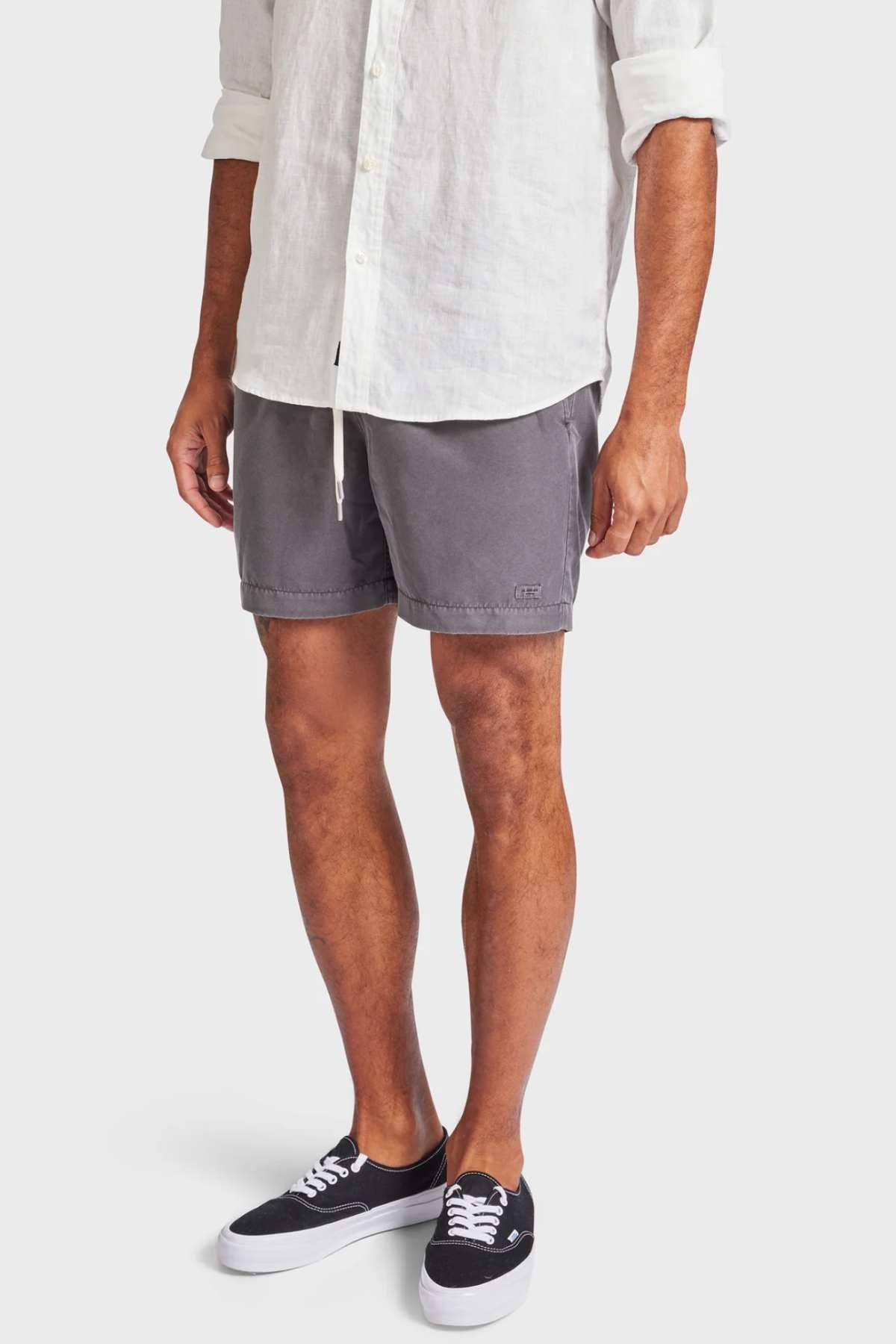 A person wearing a white long-sleeve shirt, Academy Brand Teamster Boardy tailored gray shorts made from recycled polyester, and black sneakers with white soles stands against a plain background. The untucked shirt complements the shorts' drawstring detail.