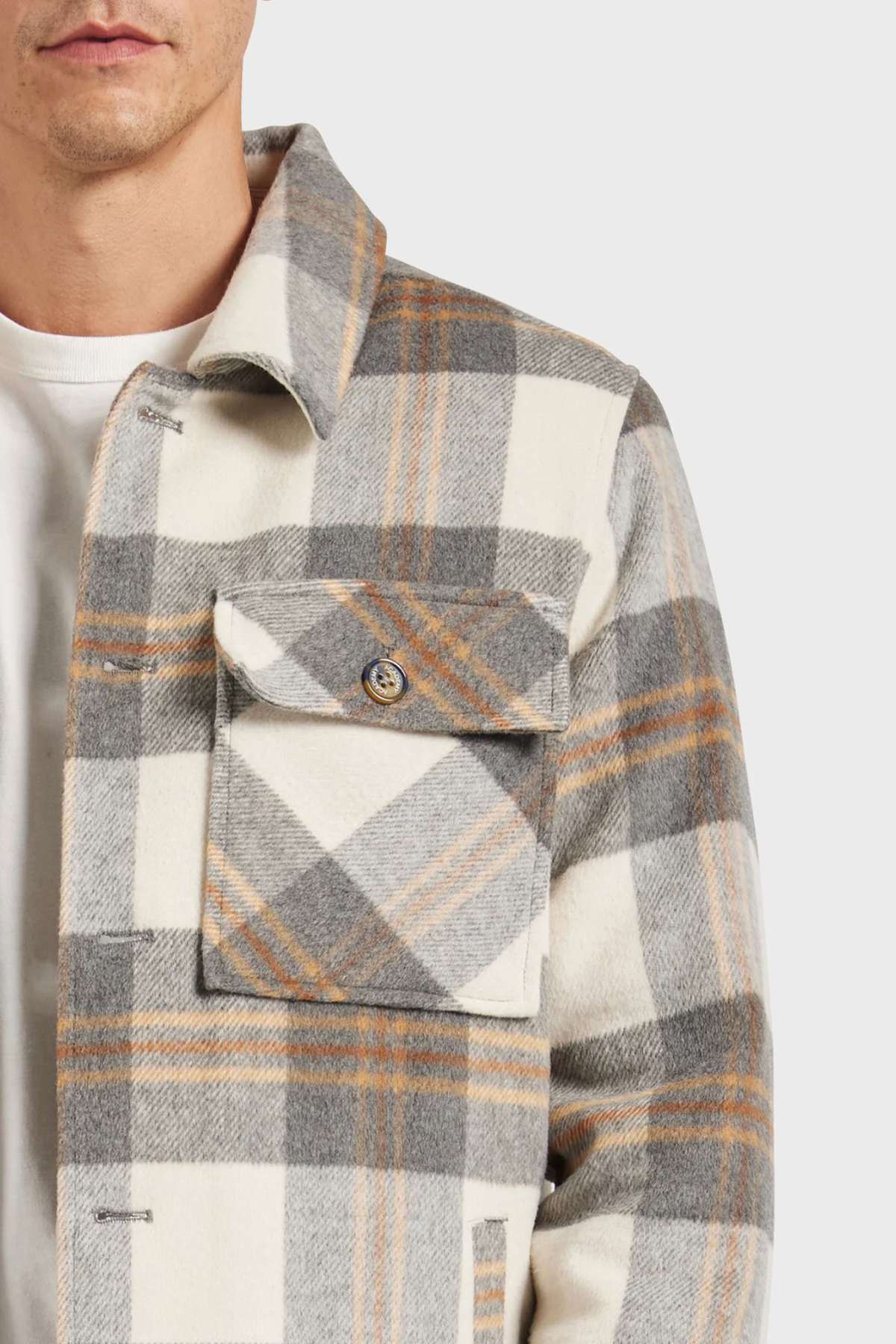 A person is wearing an Academy Brand Tulsa Jacket, a wool blend with white, gray, and light brown plaid stripes. It features a large front pocket with a button over a white shirt against a plain white background.