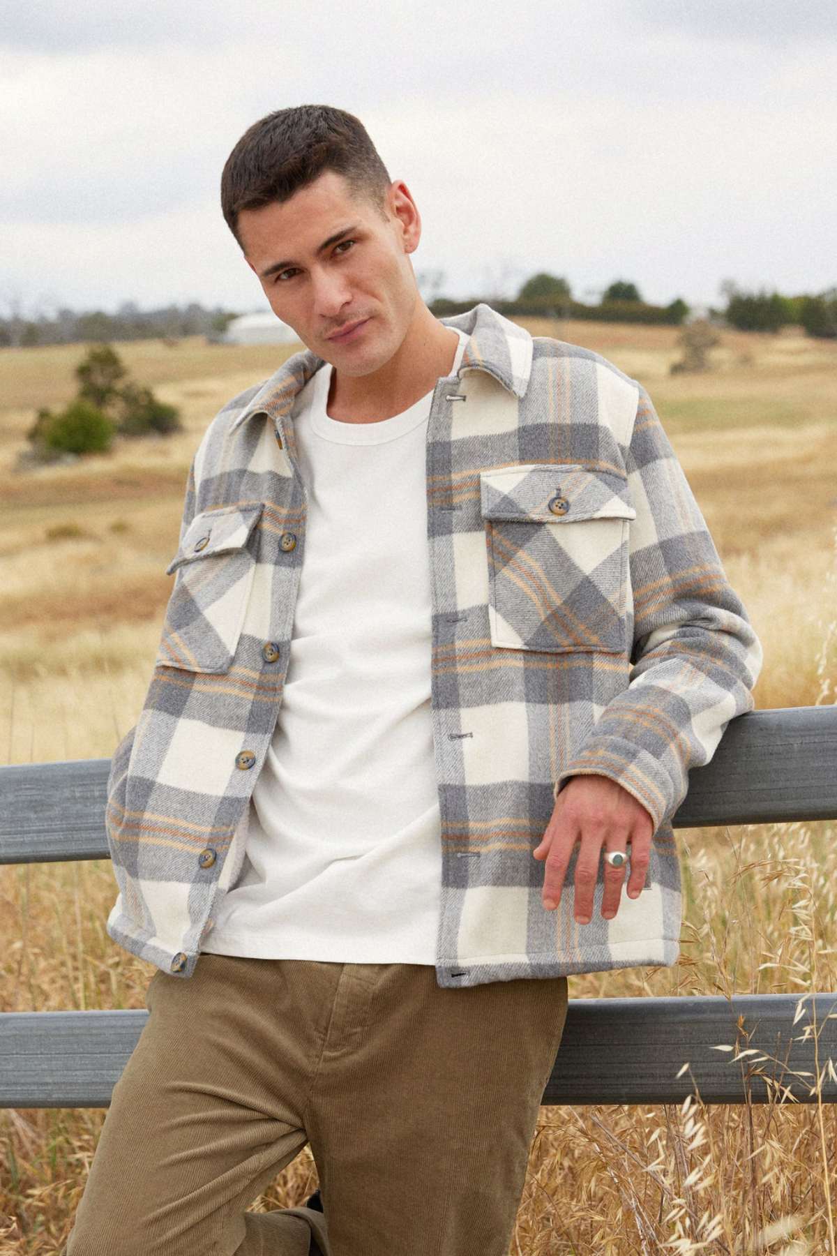 A person with short hair leans against a wooden fence in a field, donning an Academy Brand Tulsa Jacket over a white t-shirt and beige trousers. The scene is set with grass, scattered trees, and a cloudy sky.
