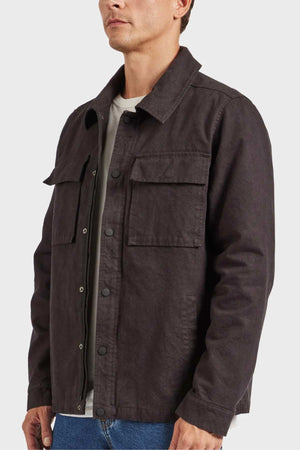 A person is wearing an Academy Brand Tulsa Jacket in dark gray. The button-up jacket, made from cotton canvas, boasts a collar, buttoned cuffs, and two chest pockets. It also features quilted lining for added warmth. Underneath the jacket is a white shirt. The person is standing against a plain white background and is visible from the chest up.