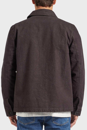 A person is standing with their back to the camera, wearing a dark-colored Academy Brand Tulsa Jacket that features long sleeves, a collar, and quilted lining for added warmth. They are also dressed in blue jeans and a white shirt that peeks out from the bottom of the jacket against a plain gray background.