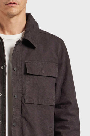 A man is partially visible, focusing on his torso and part of his face. He is wearing a dark gray Academy Brand Tulsa Jacket with quilted lining over a white shirt. The jacket, crafted from sturdy cotton canvas, features a large chest pocket with a flap. The background is plain white.