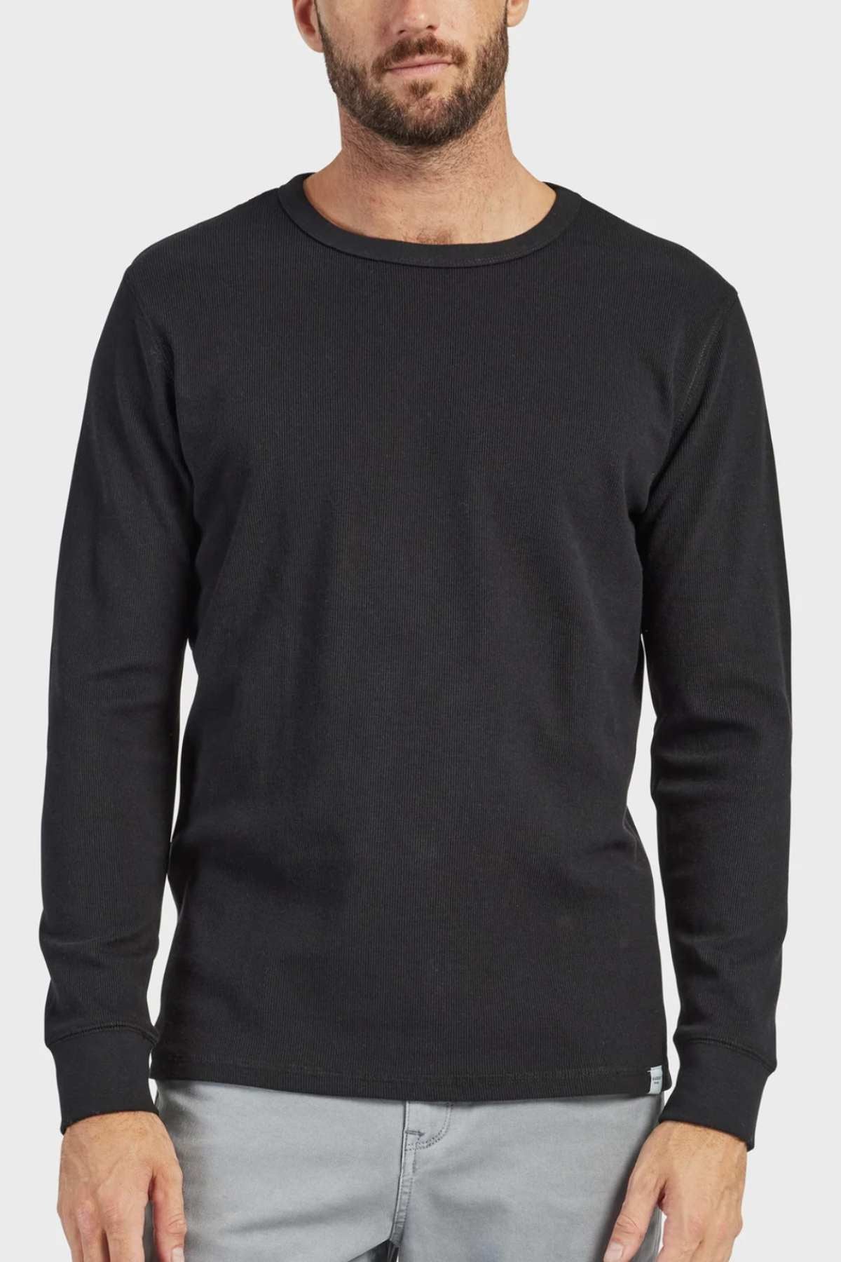 A man with a beard dons an Academy Brand Workers Crew long-sleeve, black crew neck shirt made from French rib fabric, paired with light gray pants. He stands against a plain white background.