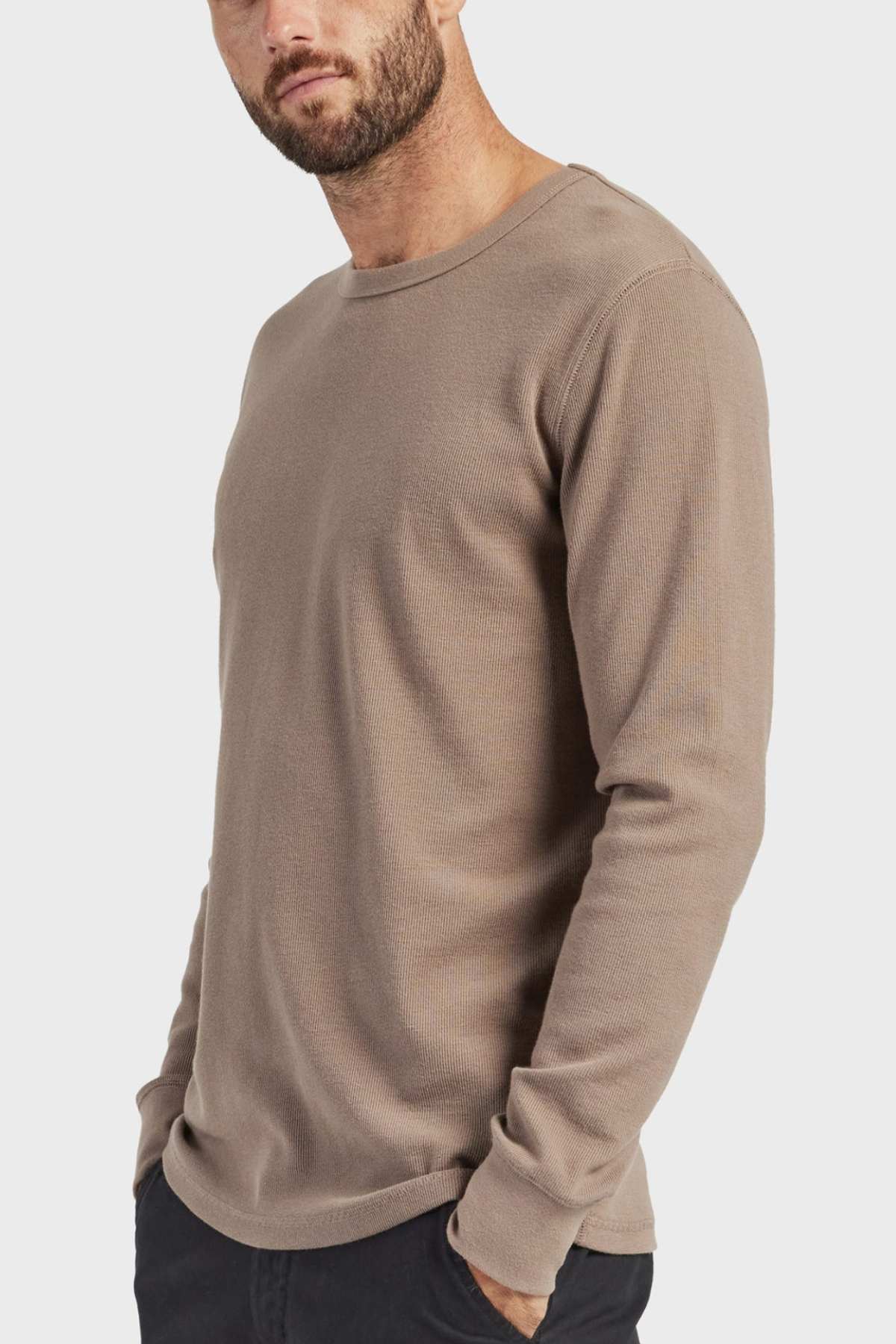 A man stands against a white background wearing a beige Academy Brand Workers Crew shirt with French rib construction and black pants. With his hands in his pockets, he gazes to the side, highlighting the shirt's durable design.